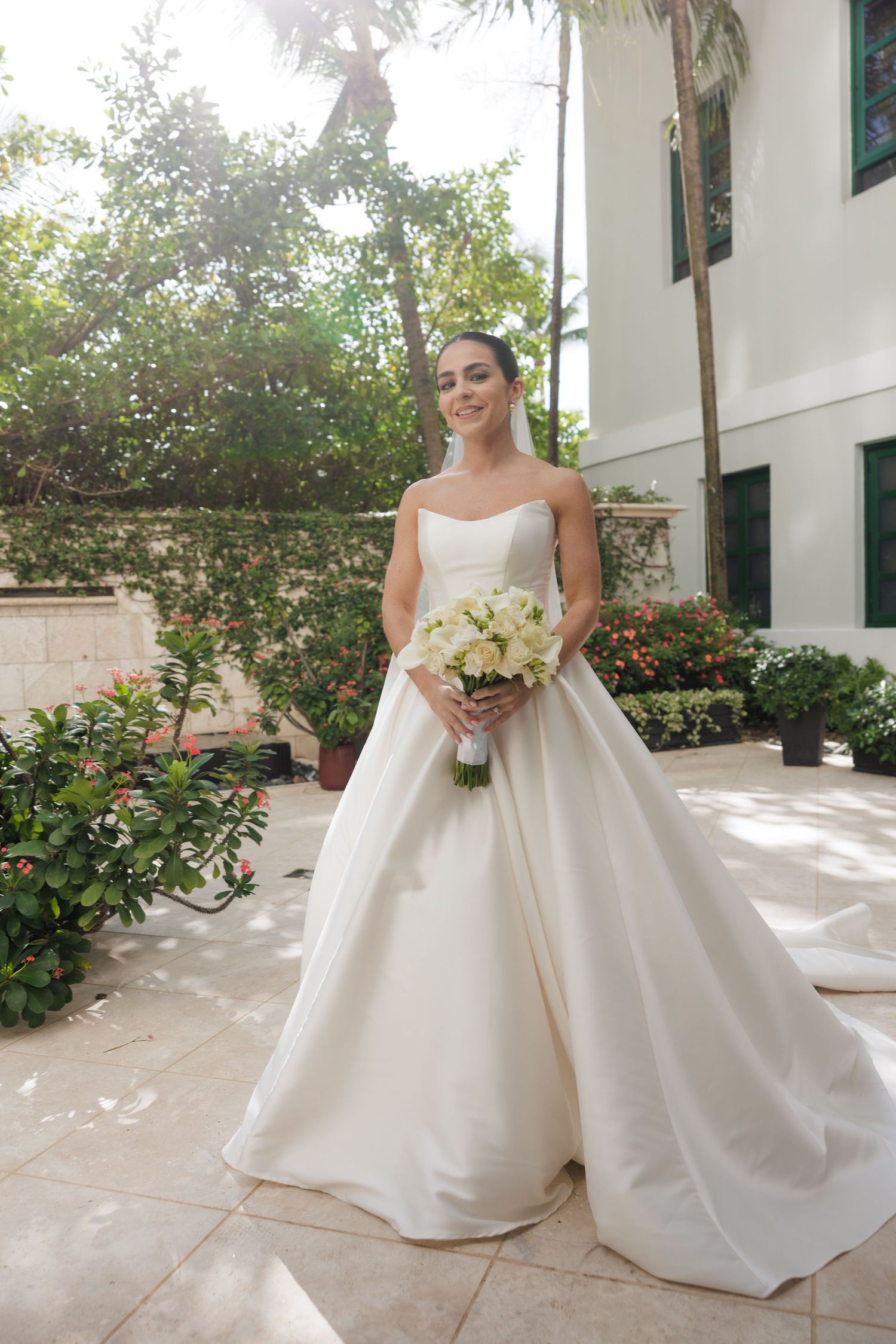 Bride in classic white ballgown poses in sunlit courtyard with flowering bushes.