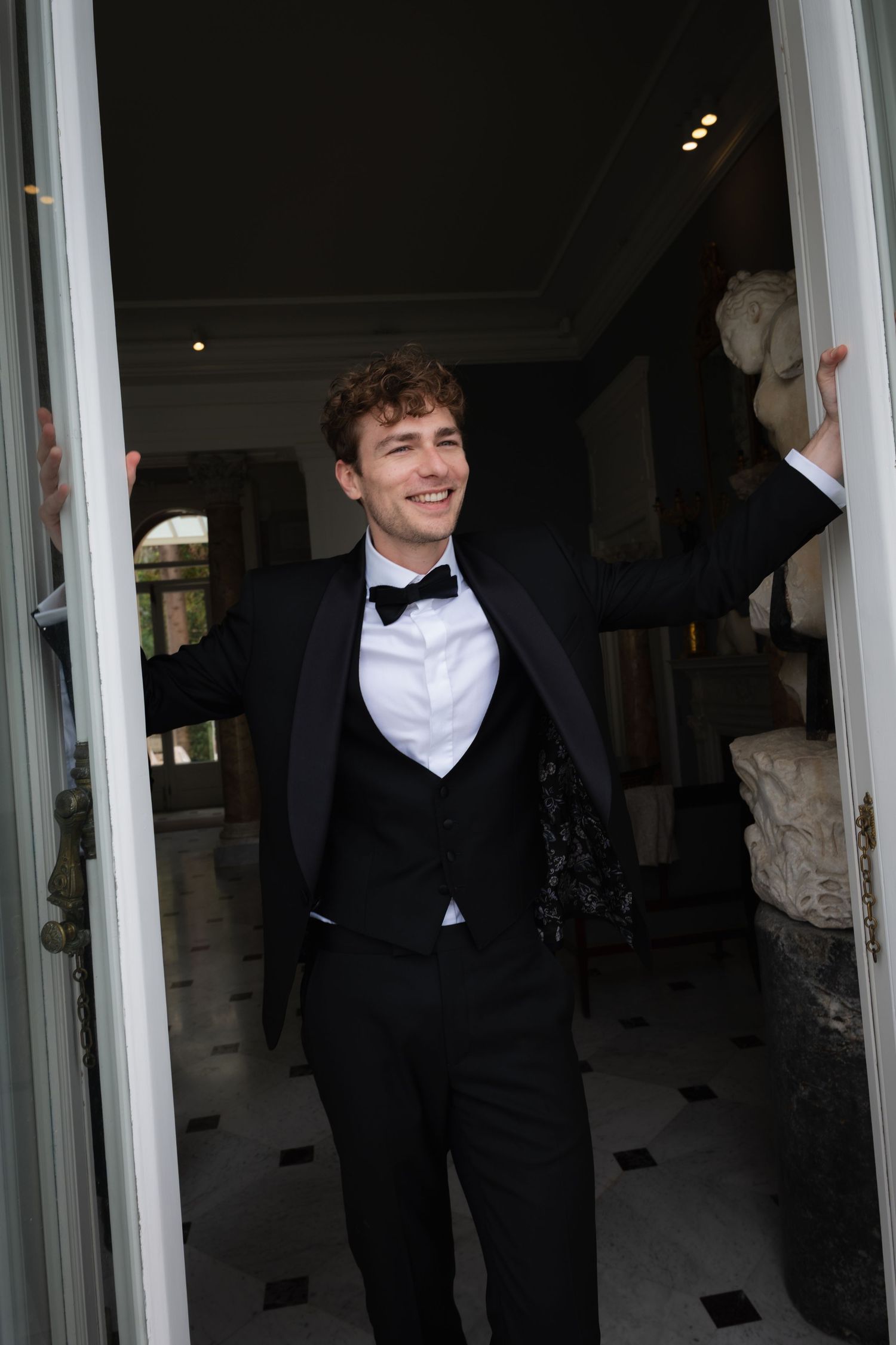 Young man in black tuxedo with bow tie smiling while standing in elegant doorway with classical sculptures.
