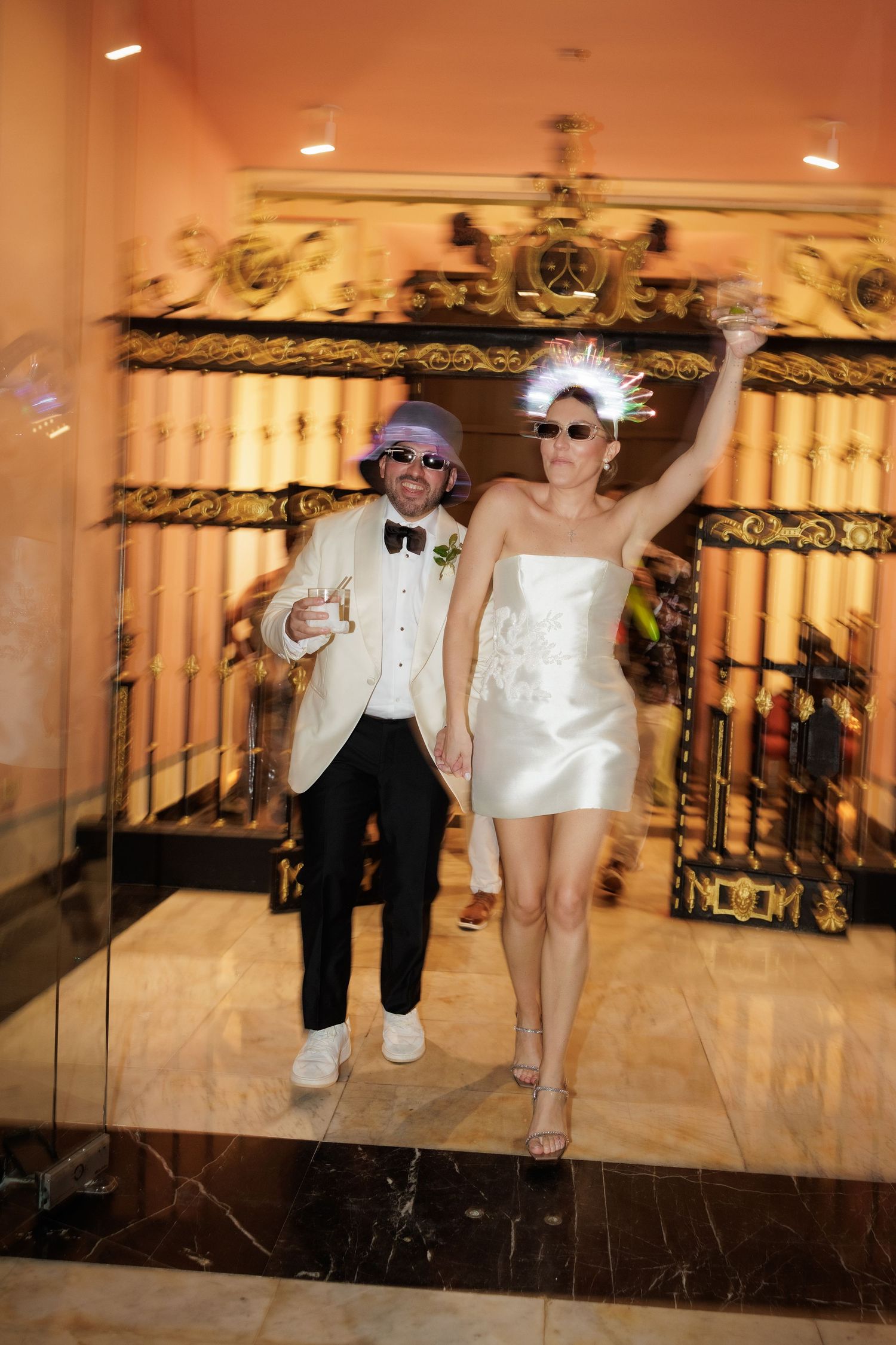 Two people wearing sunglasses and formal attire dance and pose in a hotel hallway with ornate gold railings.
