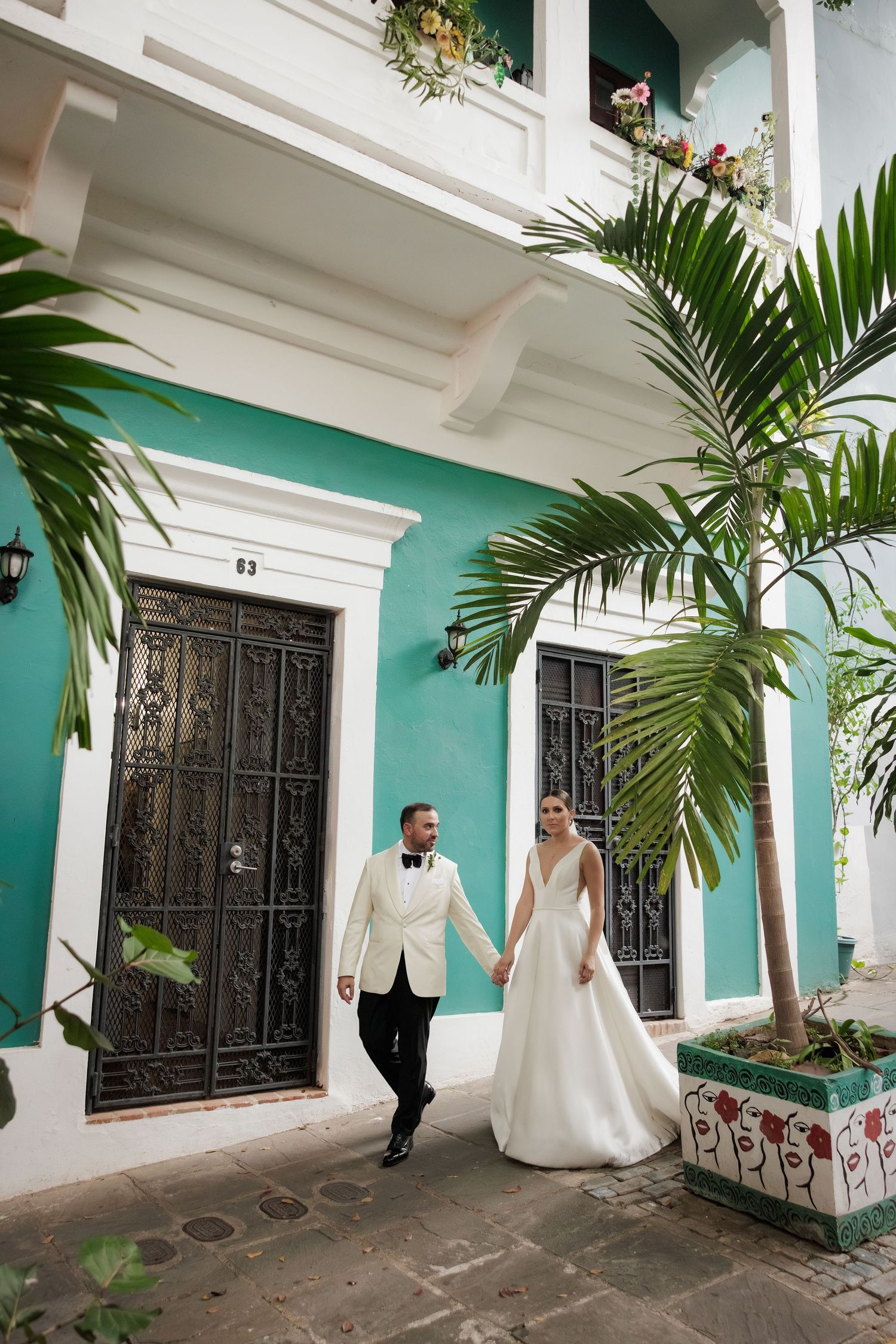 A couple in wedding attire walks along a turquoise-walled colonial building with palm trees and ornate windows.