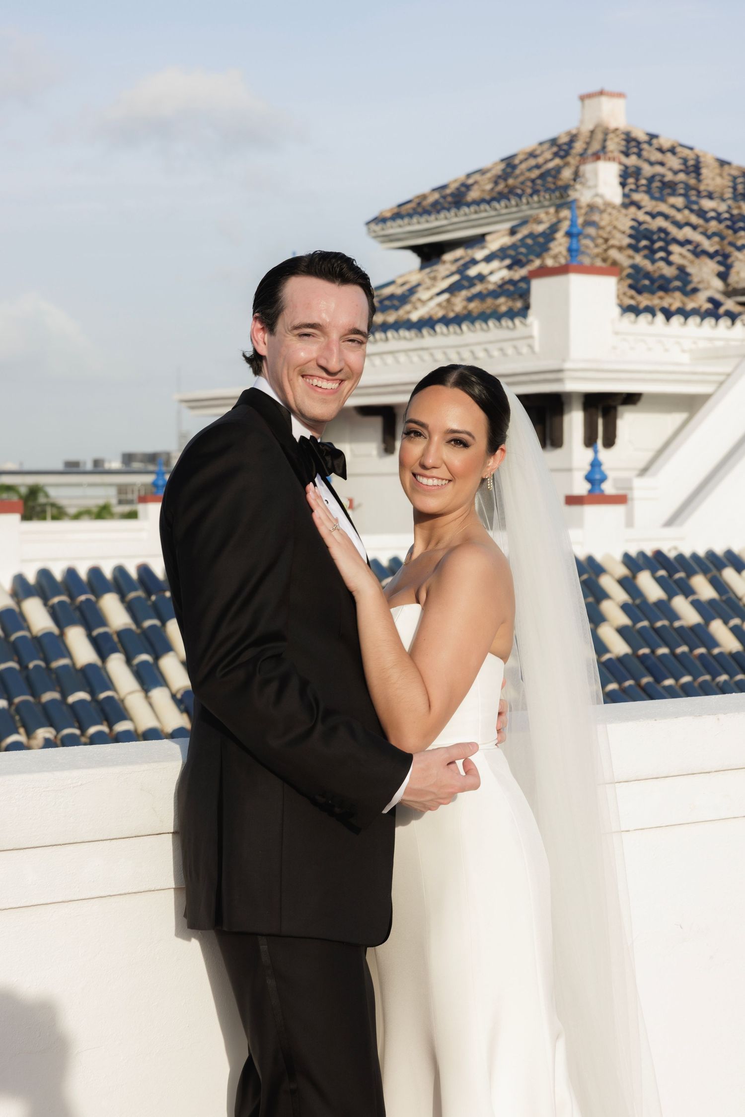 A bride and groom share an intimate moment on a rooftop terrace with ornate architecture and blue seating in view.