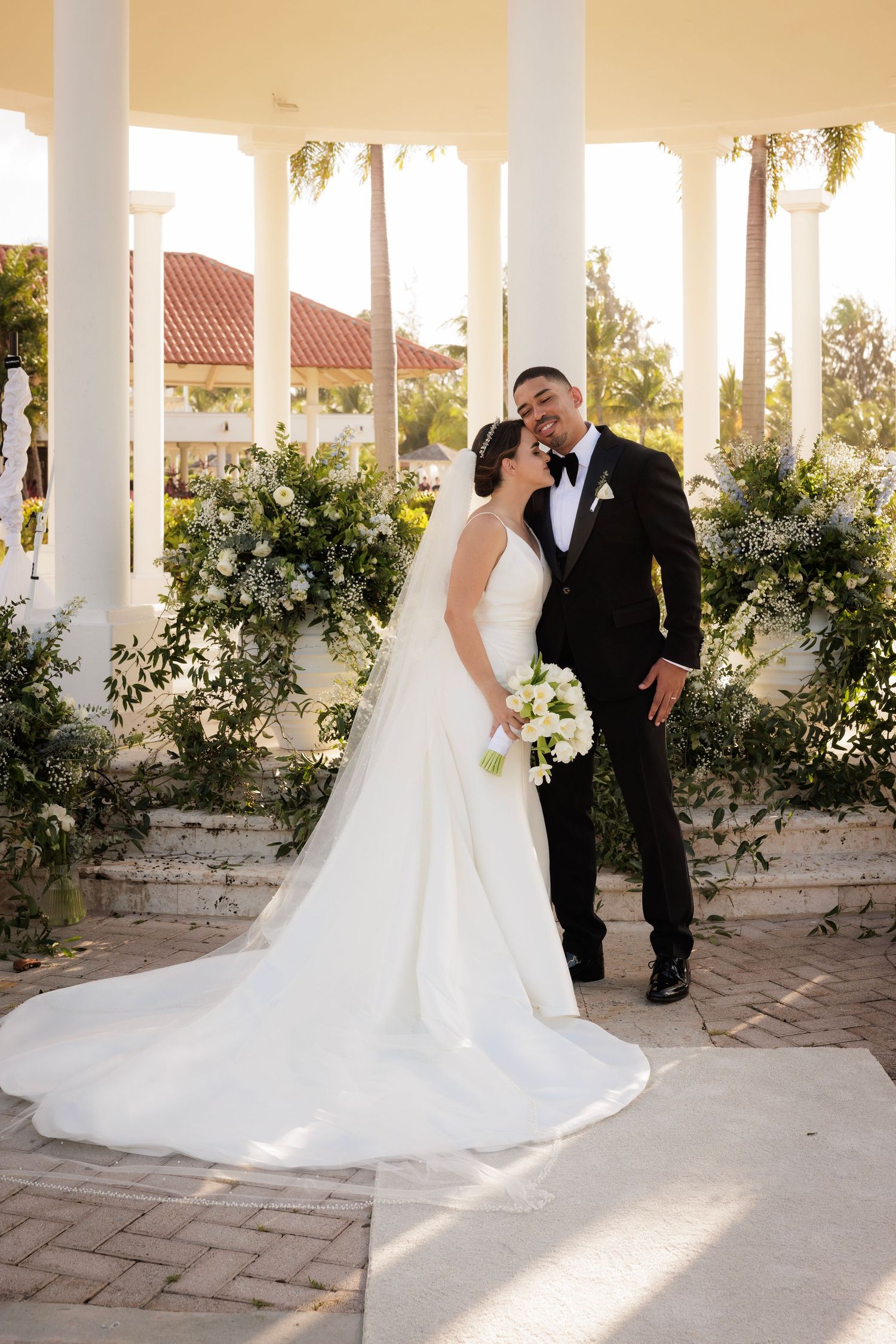 Bride and groom share intimate moments under a white pillared walkway with trailing vines at a garden wedding venue.