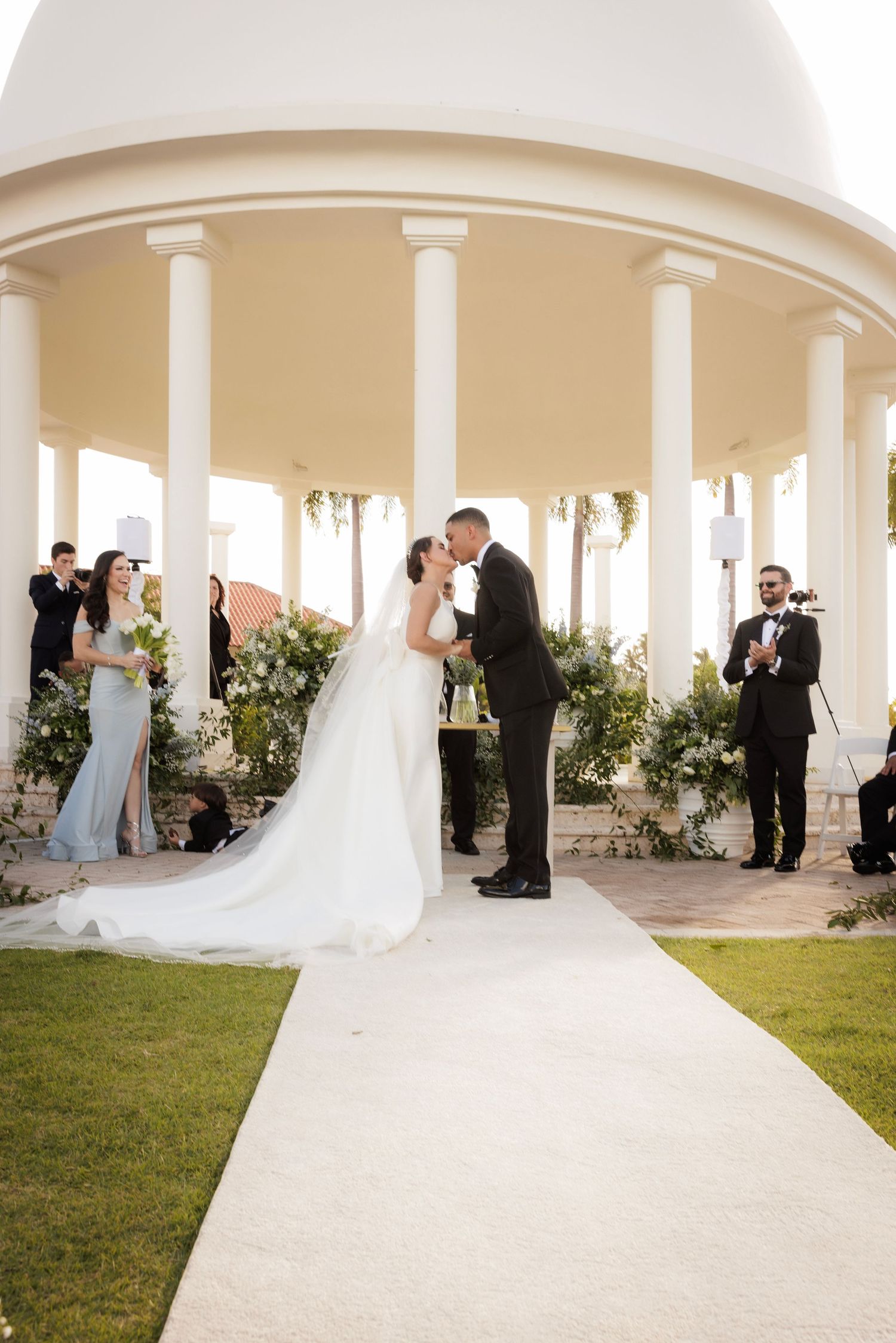 Elegant wedding ceremony takes place under white pillared colonnade with decorative greenery and guests on the lawn.