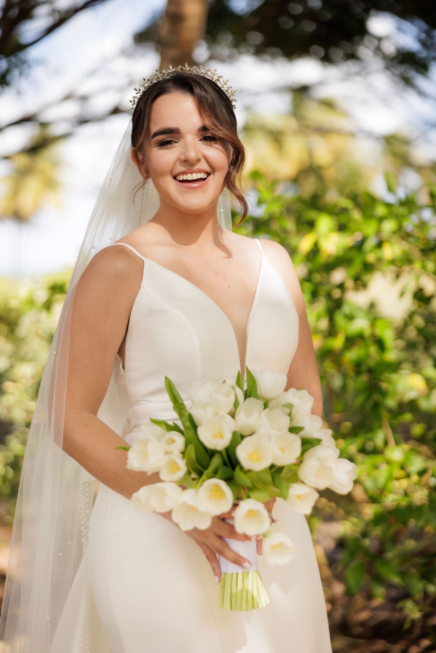 A bride in a white wedding dress holds a bouquet of white flowers while posing in a natural outdoor setting.