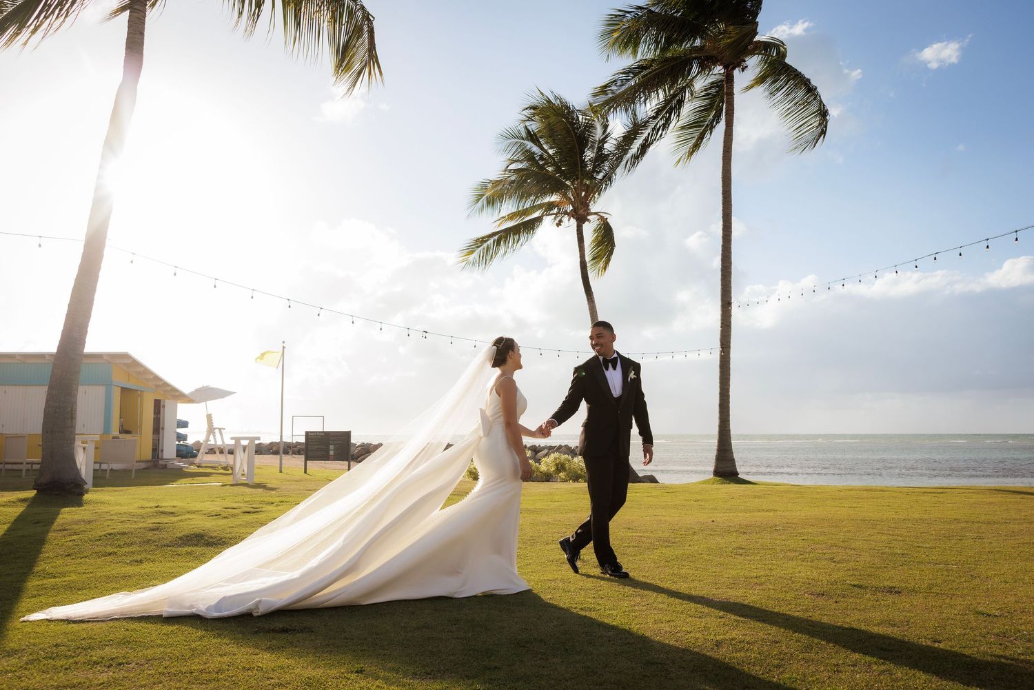 A long wedding veil flows gracefully in the breeze against a backdrop of palm trees and ocean at a tropical beach destination.