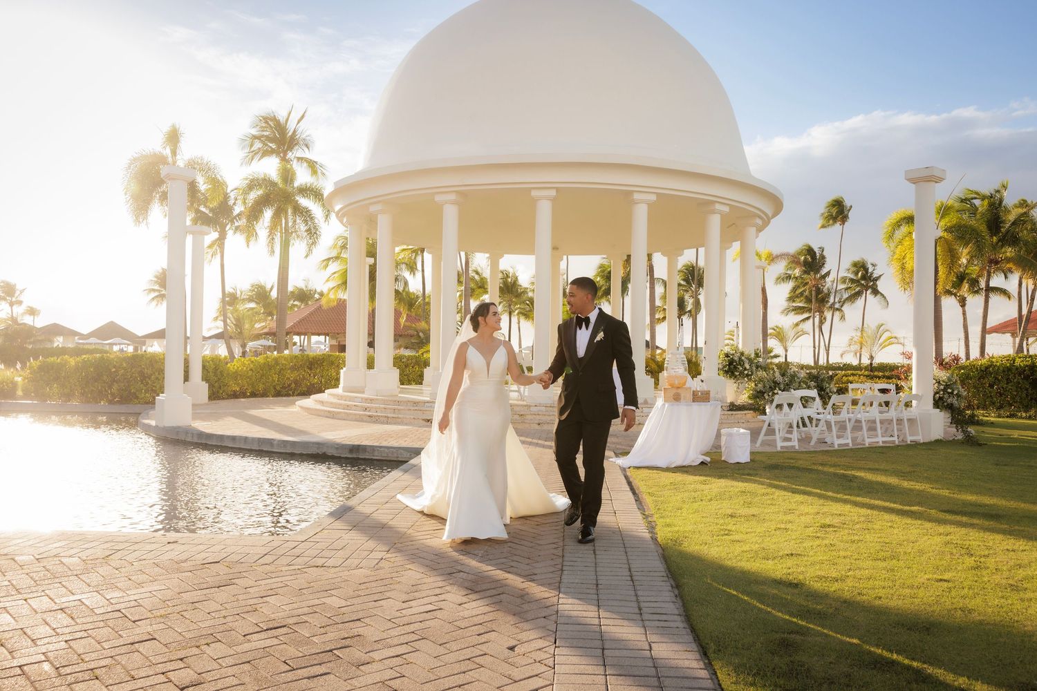 A scenic outdoor beach wedding ceremony with white archway and chairs set up along the oceanfront under palm trees.