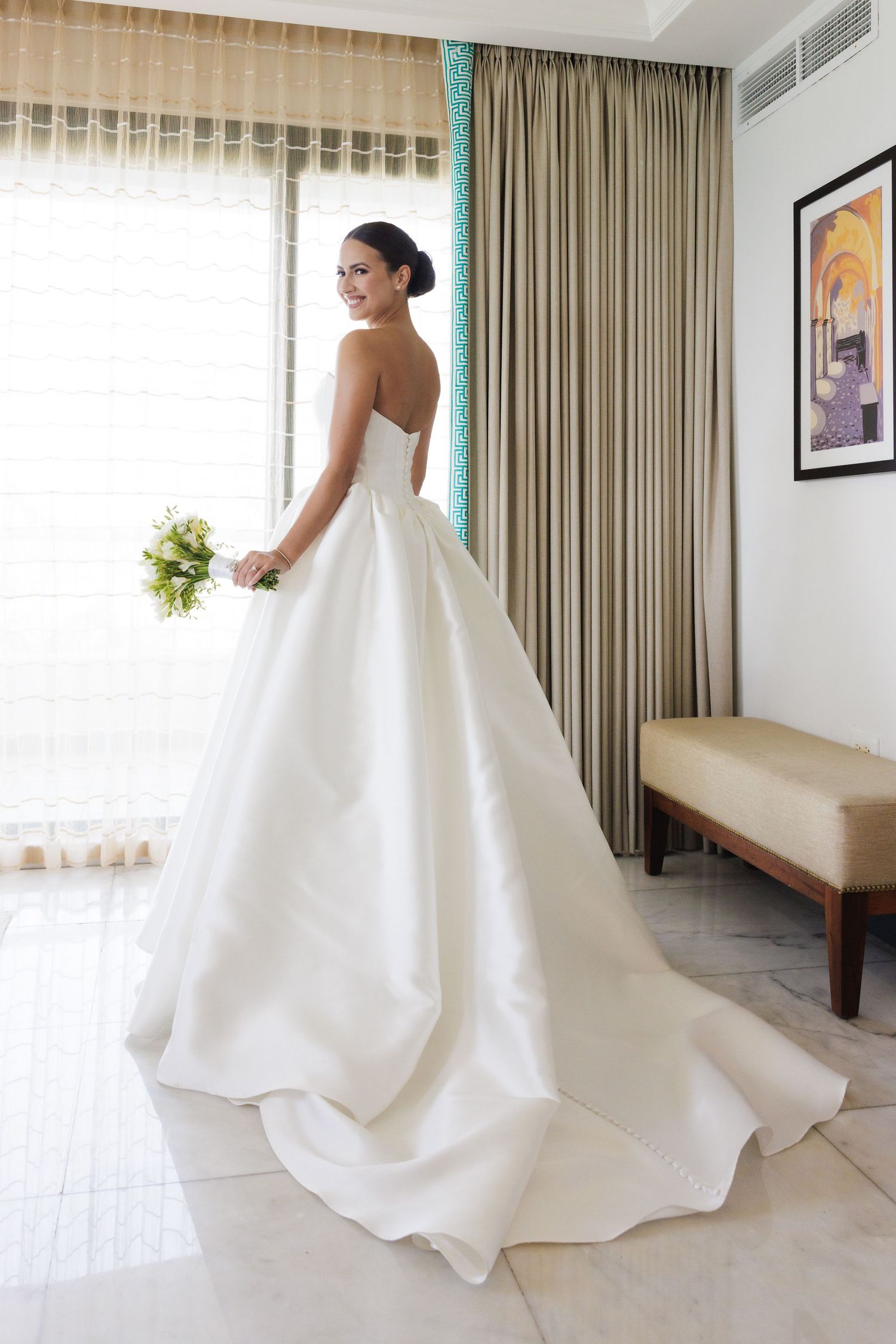 Elegant white wedding gown with dramatic train flows across a modern hotel room interior.