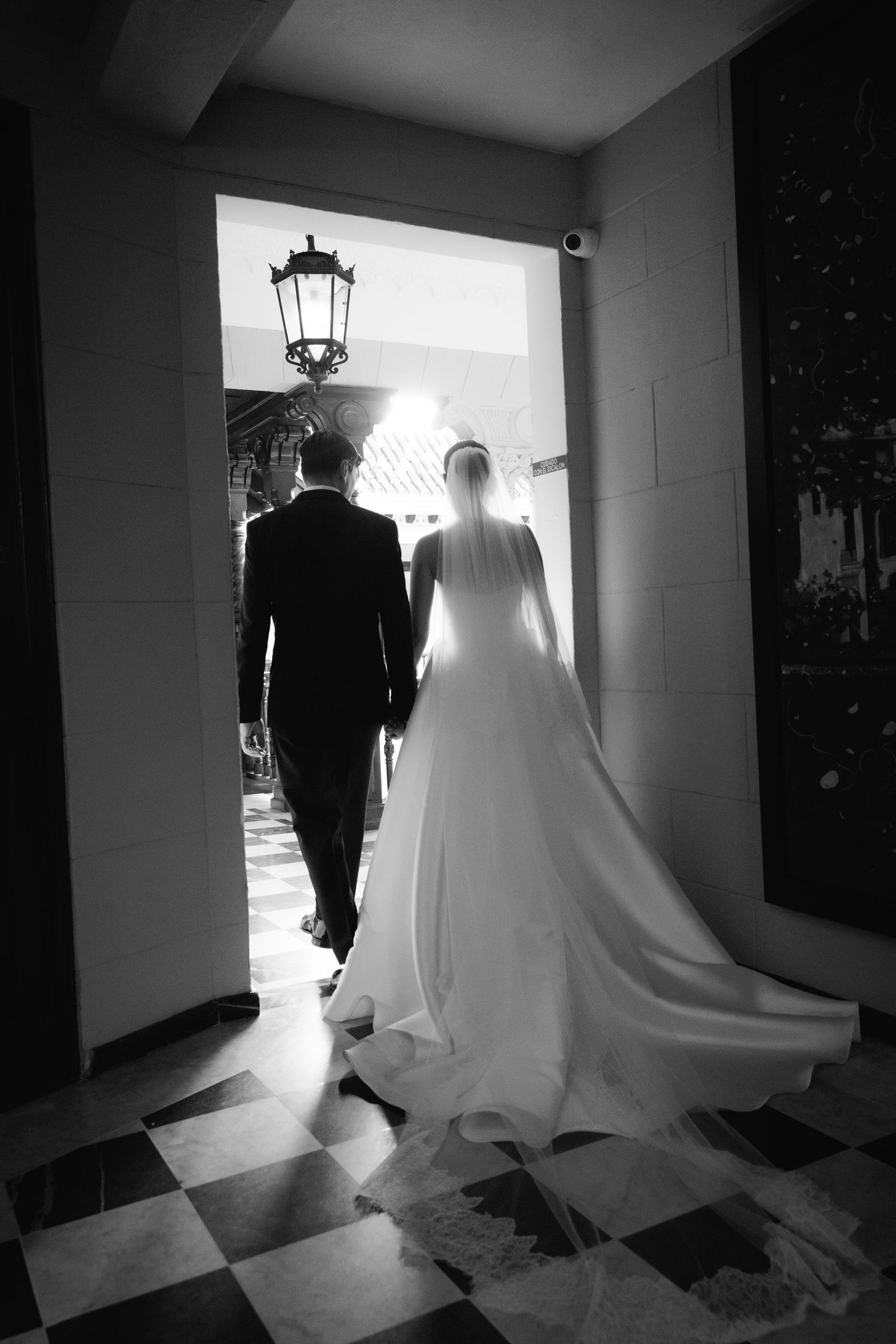 A dramatic black and white silhouette of a wedding dress and suit in a hallway with bright backlighting.
