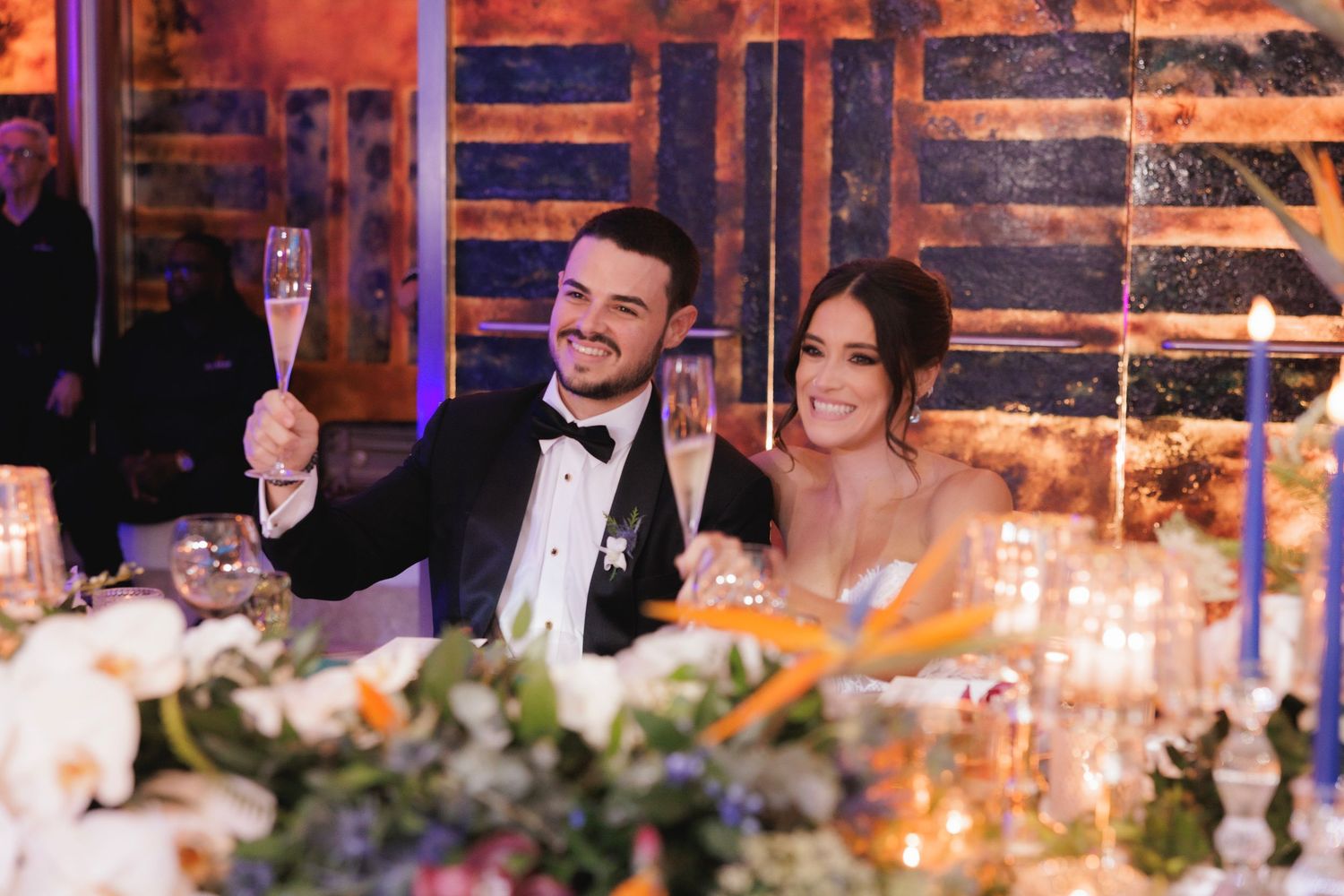 A candlelit wedding reception table decorated with floral centerpieces and glowing tapers in an elegant rustic venue.