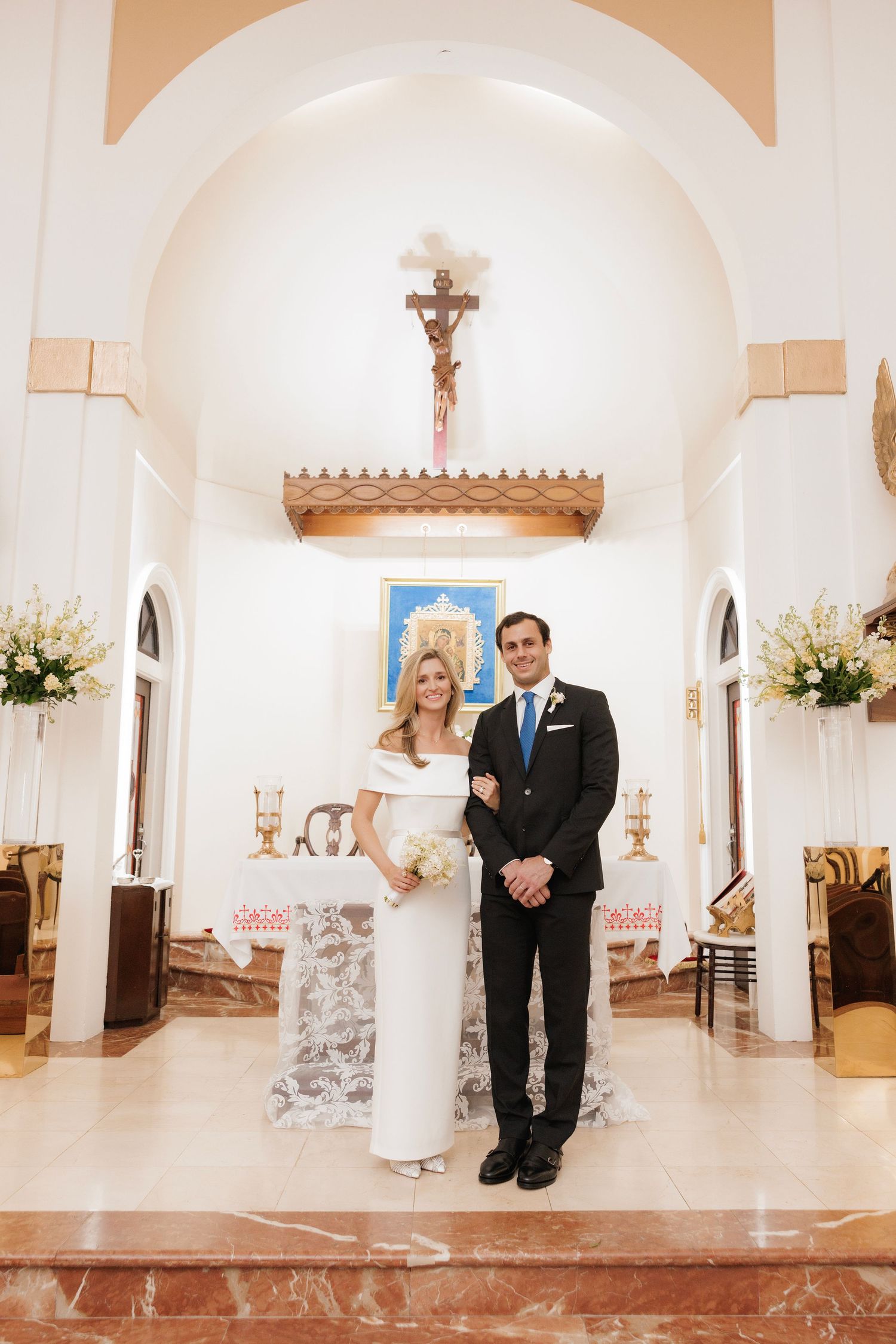 A newly married couple stands at the altar of a traditional Catholic church with white walls and floral arrangements.