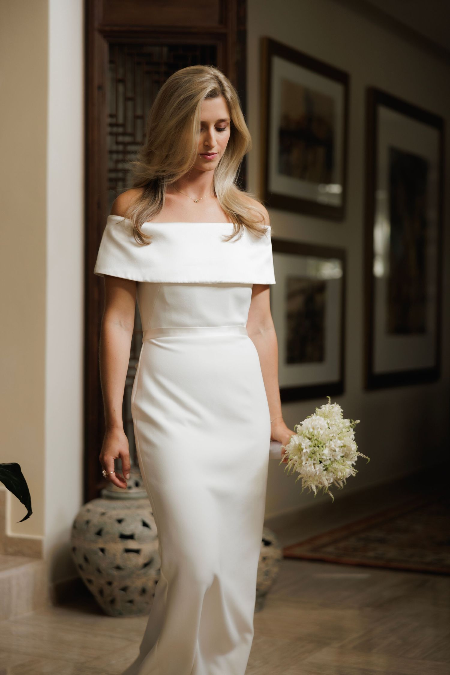 A bride in an elegant off-shoulder white wedding gown holds a cream-colored bouquet in a hallway with framed artwork.