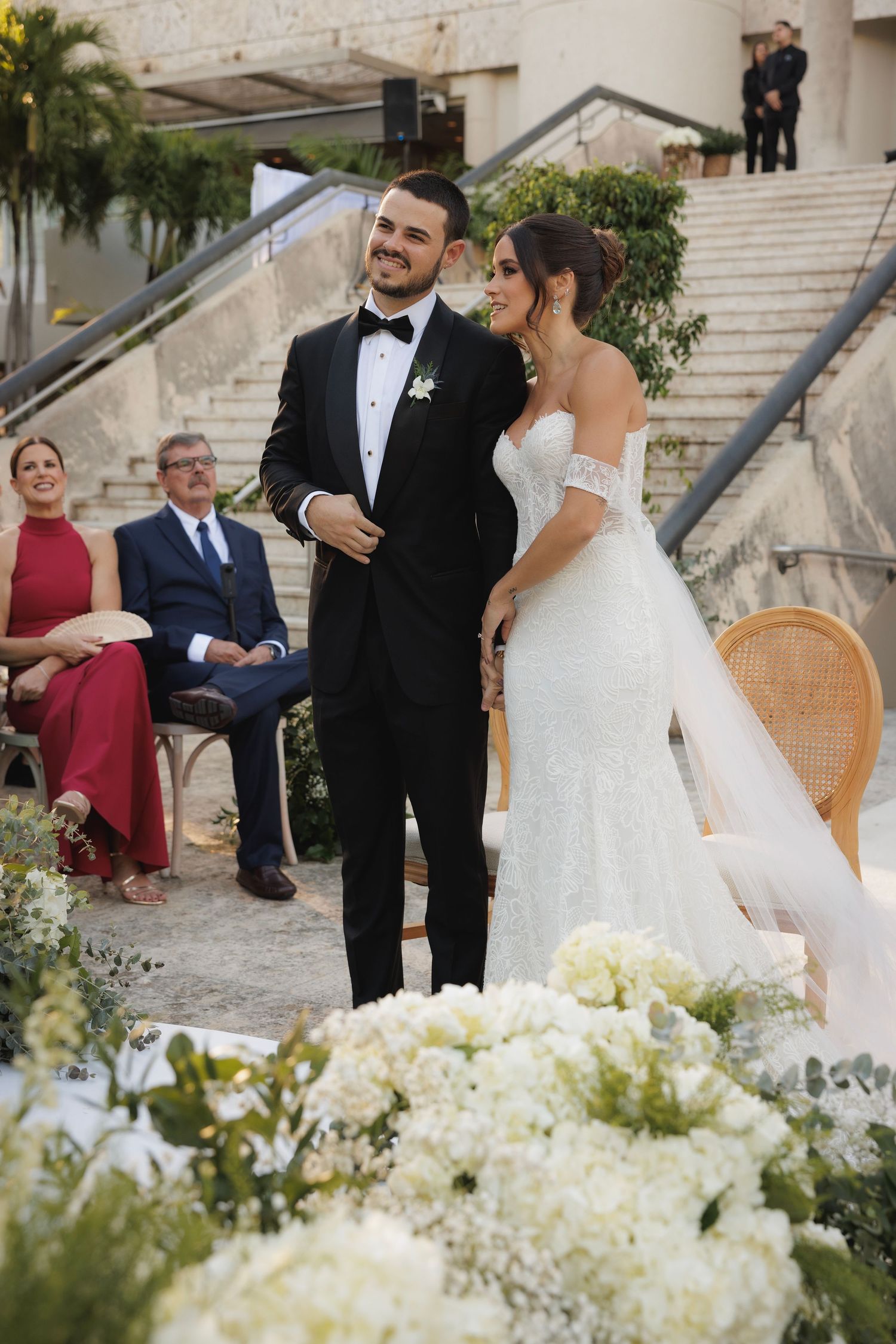 A wedding couple shares an intimate moment outdoors with white floral decorations and elegant seating.