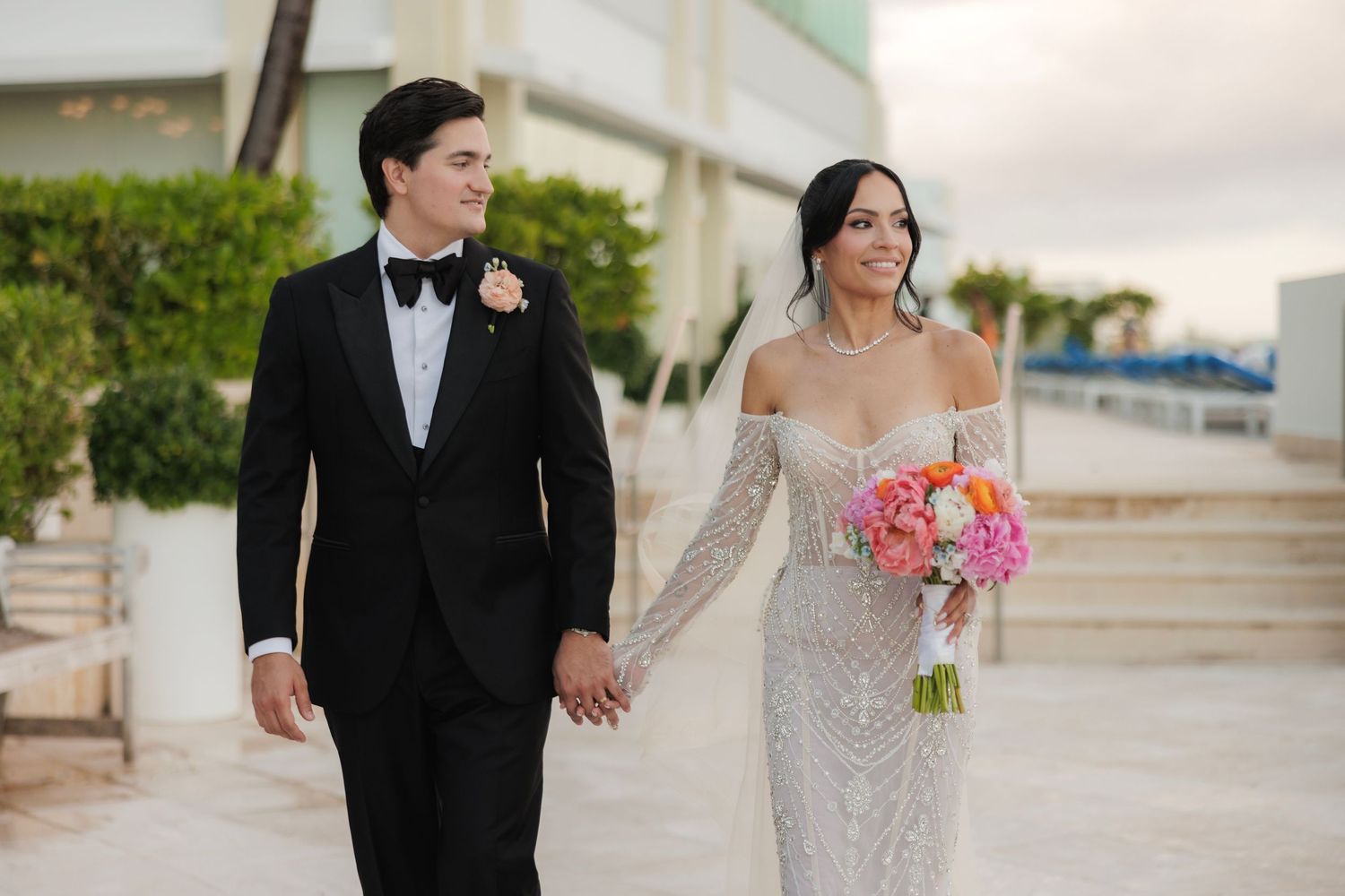 A bride in a white off-shoulder gown holds a pink bouquet while walking with her groom in a black tuxedo.