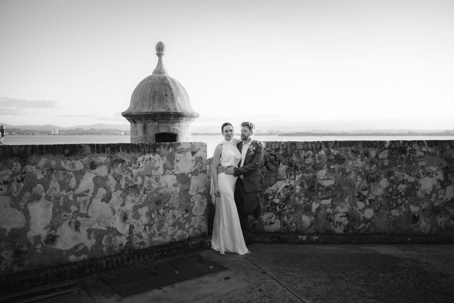 A couple stands in an elegant black and white moment against a stone wall with domed turret at El Morro fortress in Puerto Rico.