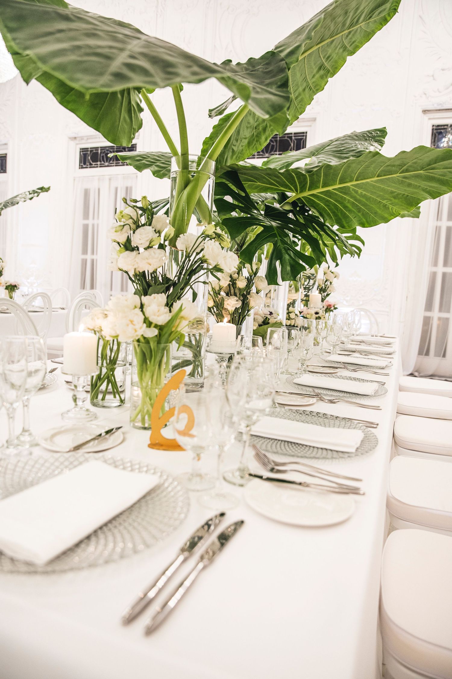Elegant white table setting with tropical leaves and glassware arranged in an open gallery space.