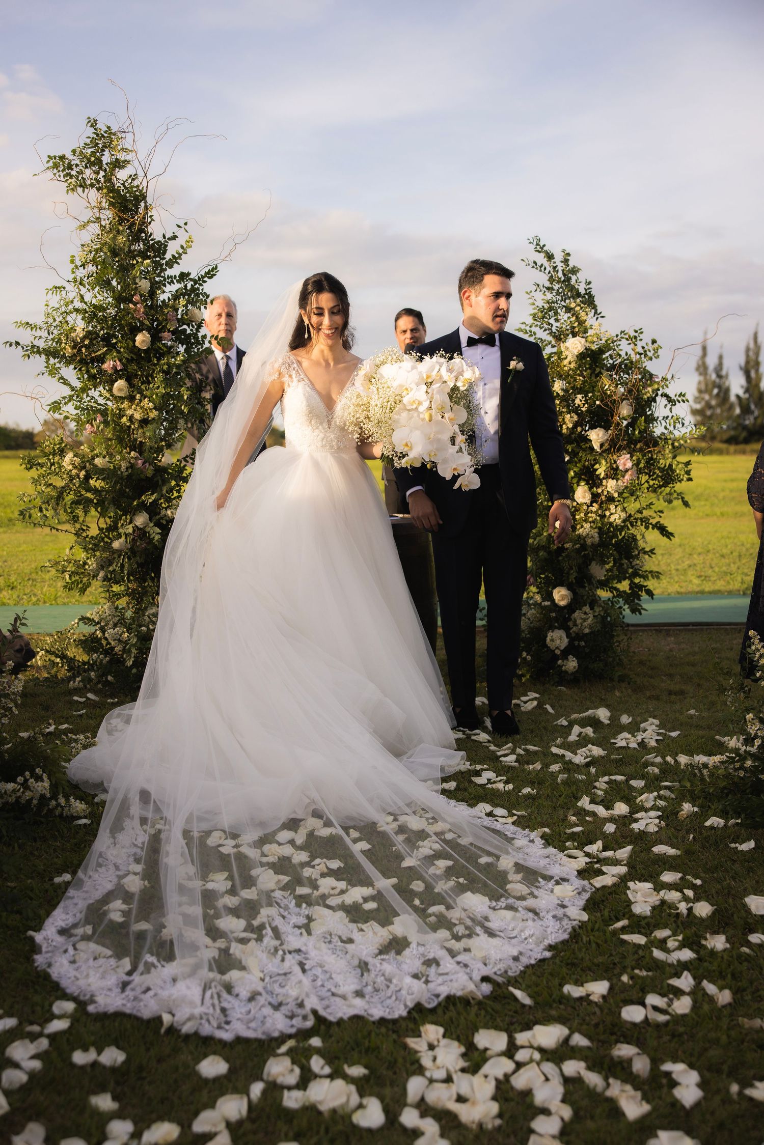 A wedding couple stands in a garden setting with scattered white rose petals on the grass during golden hour.