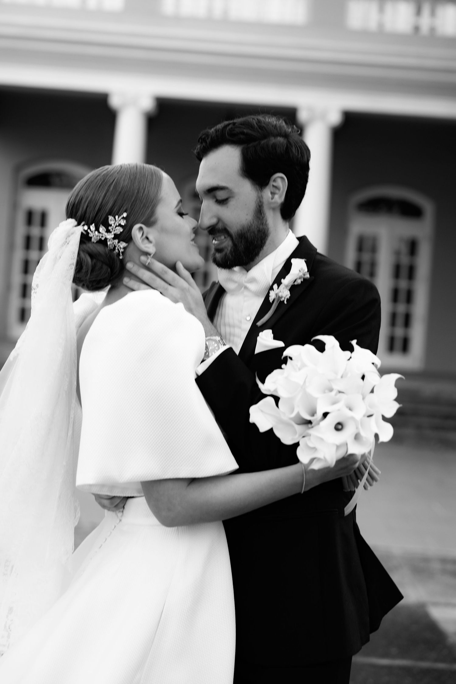 A bride and groom share a romantic moment in black and white, with her holding a white lily bouquet.