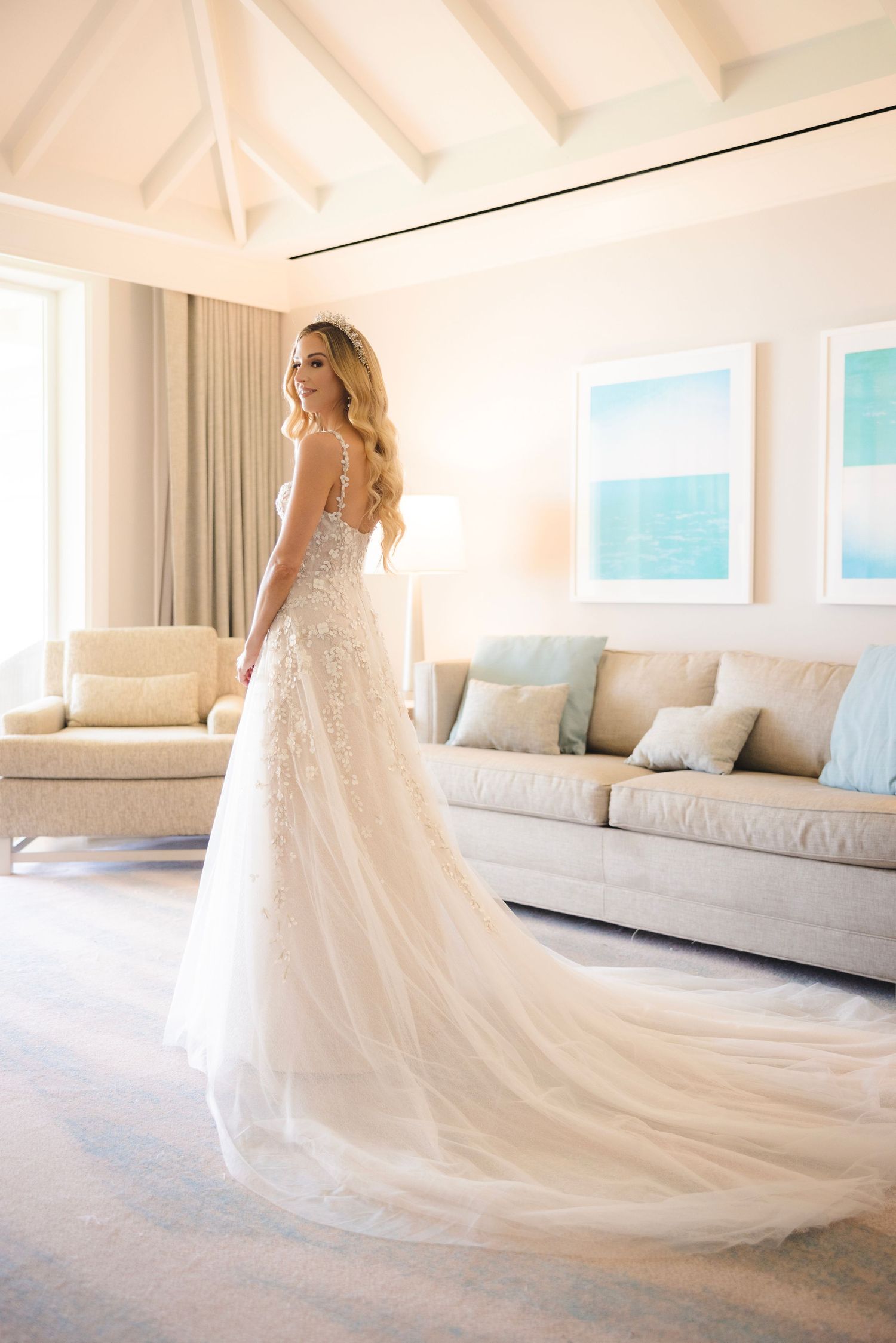 A bride in a flowing lace wedding gown stands in an elegant white room with coastal artwork and beige furniture.