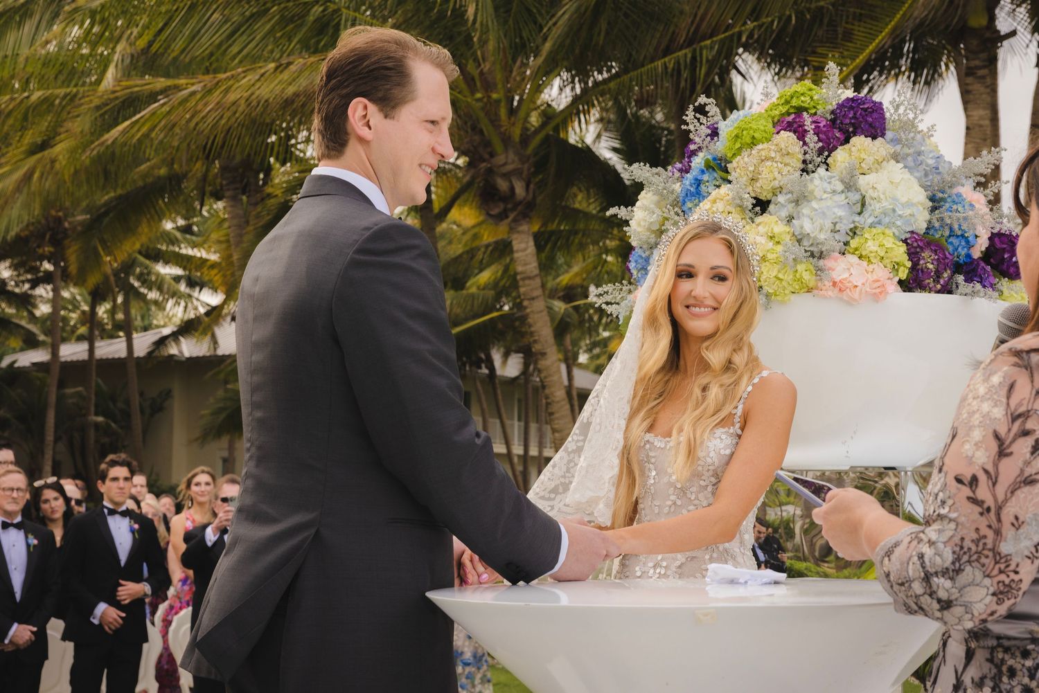 A couple shares a joyful moment during their outdoor wedding ceremony beneath palm trees with colorful floral arrangements.