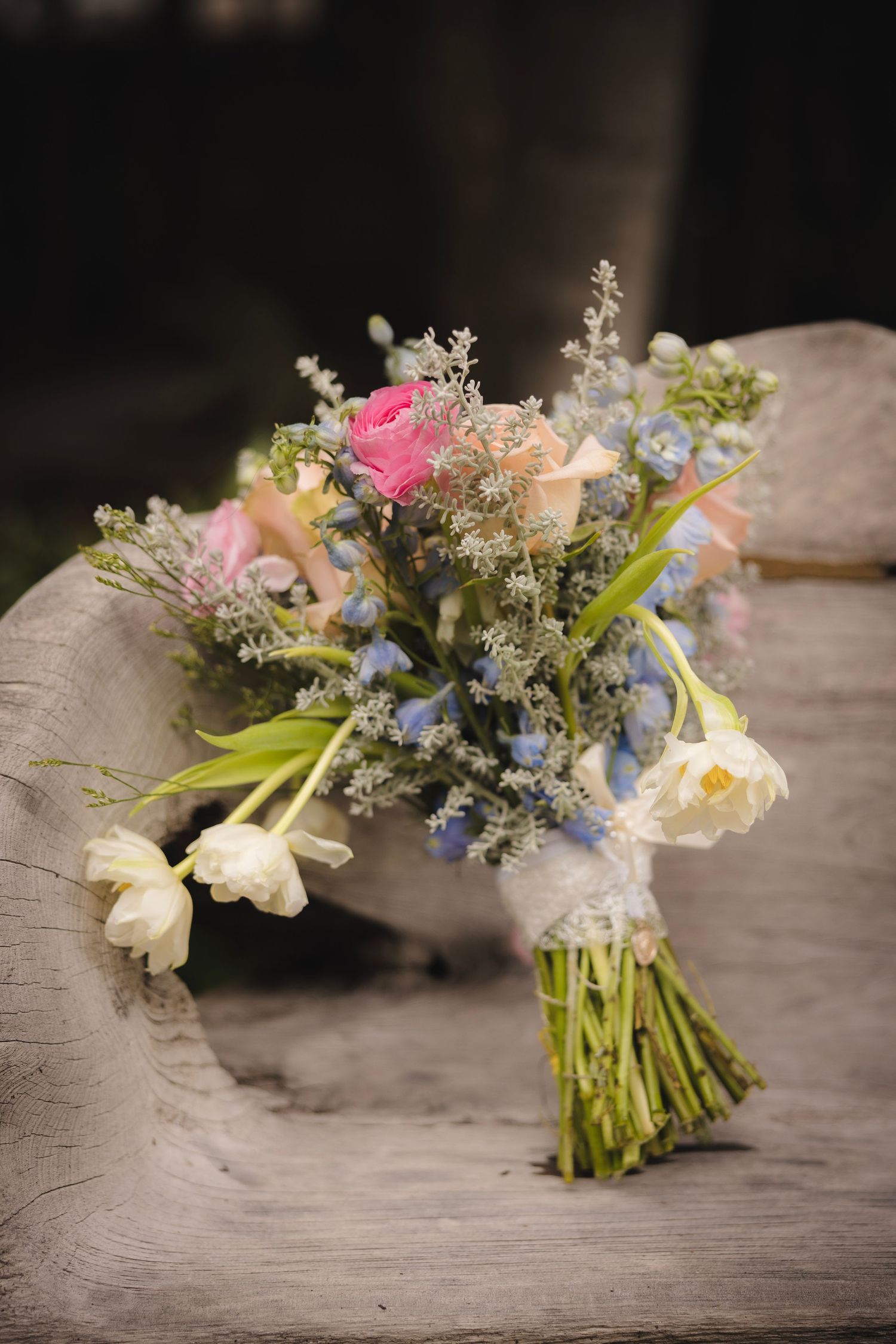 A rustic bridal bouquet with pink roses, blue accents, and delicate white flowers tied together on a wooden surface.