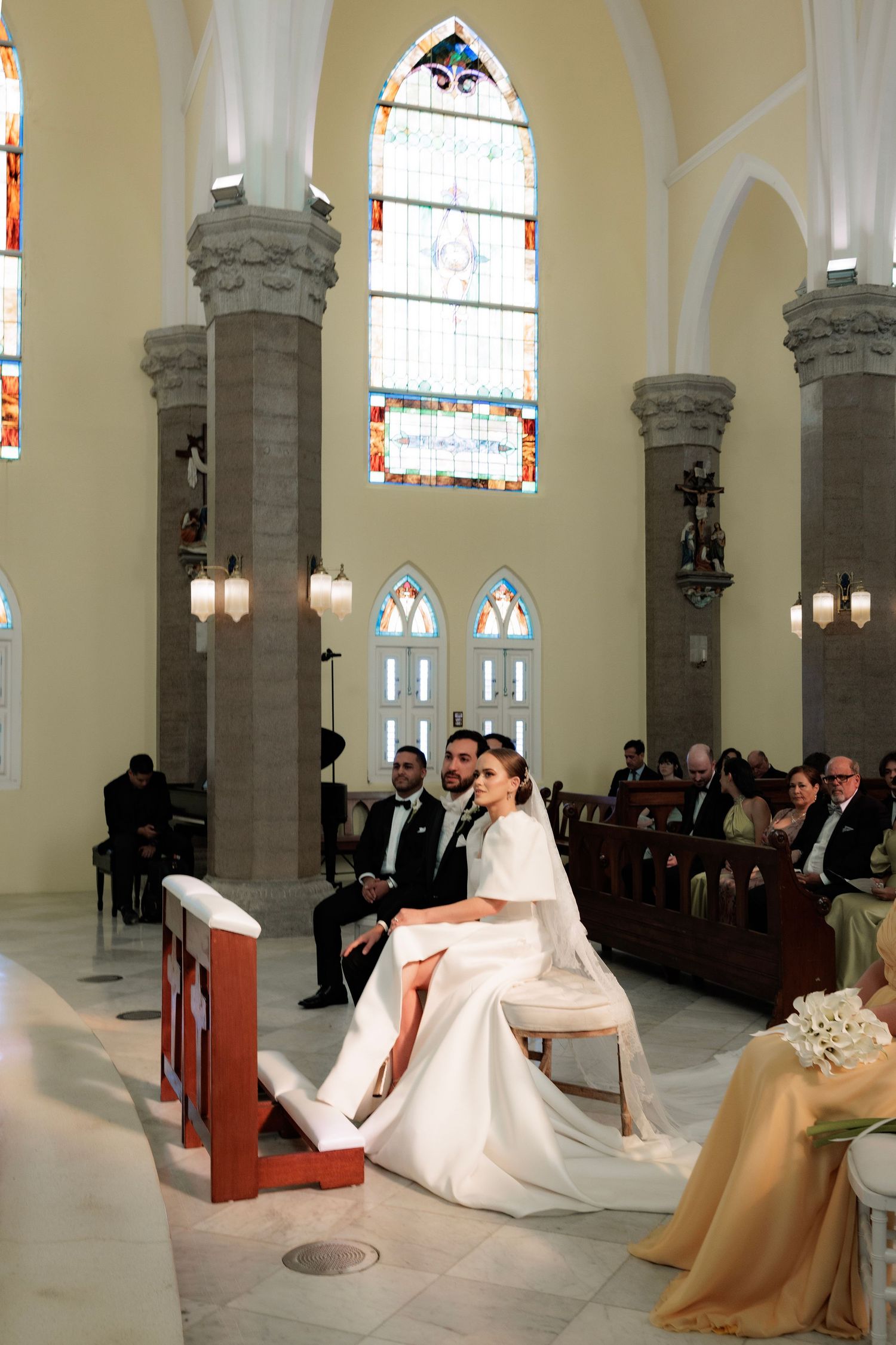 A bride in a flowing white gown kneels in prayer at a traditional church altar during a wedding ceremony.