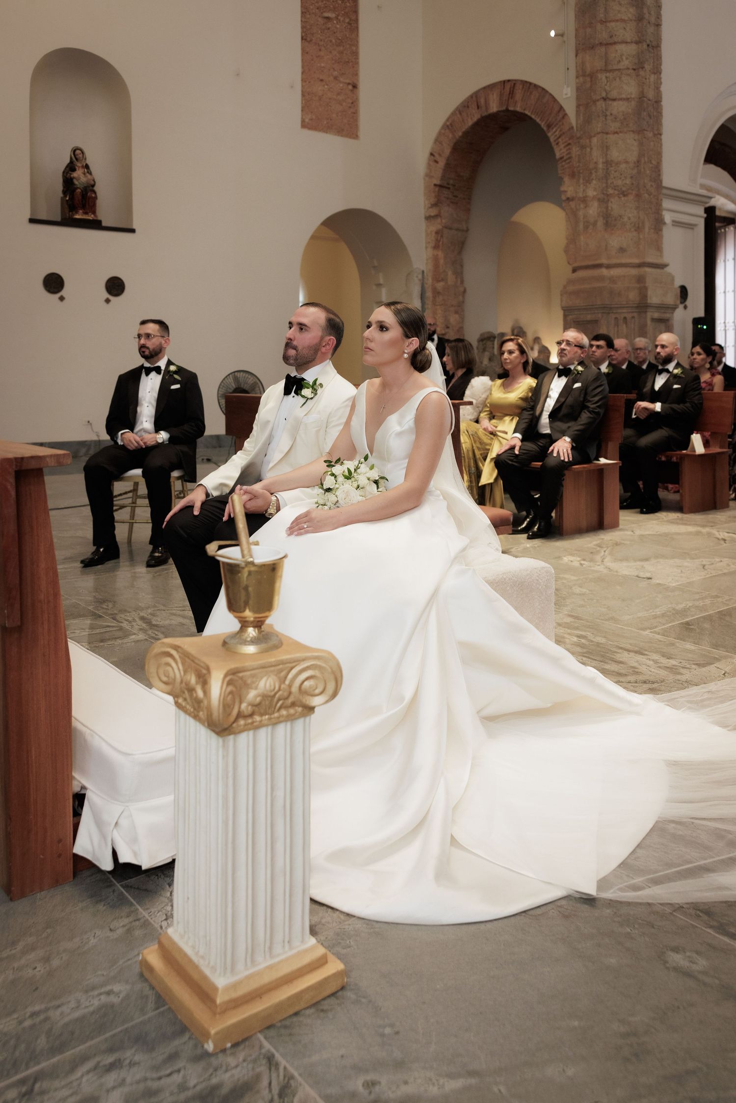 A couple sits during a wedding ceremony in a historic church with stone archways and brick details.