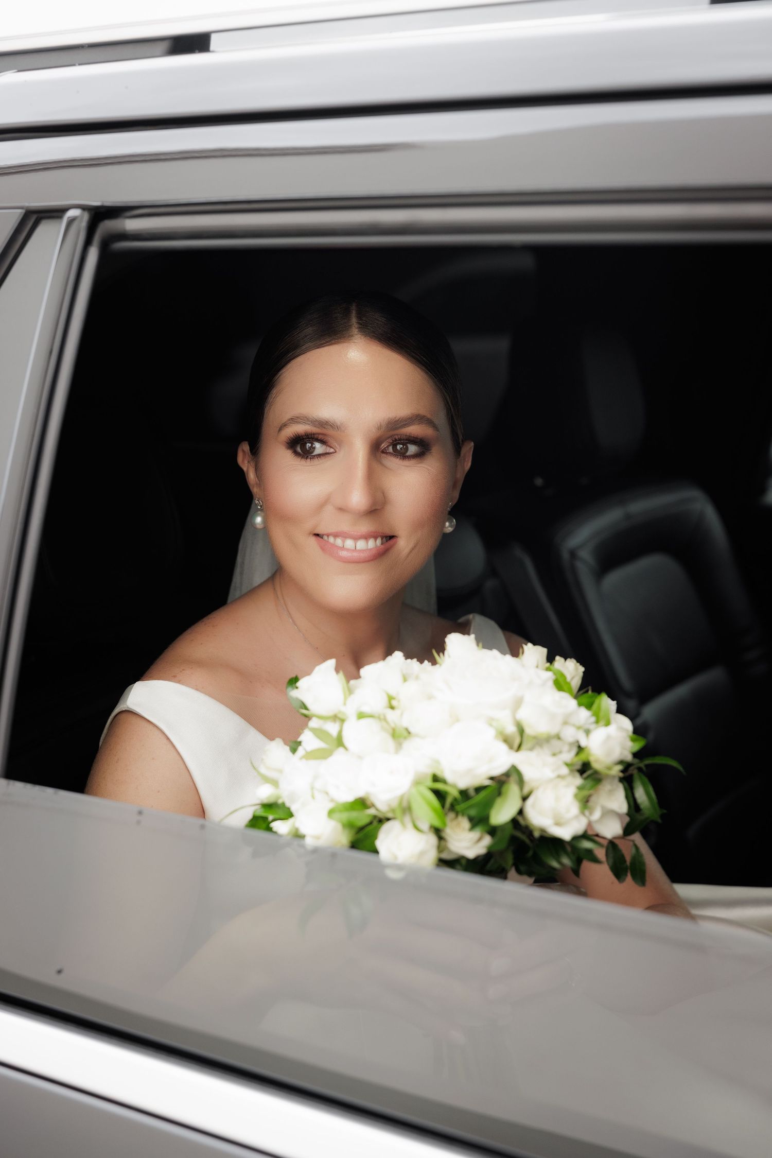 Bride with white flowers smiles from inside a wedding car window.