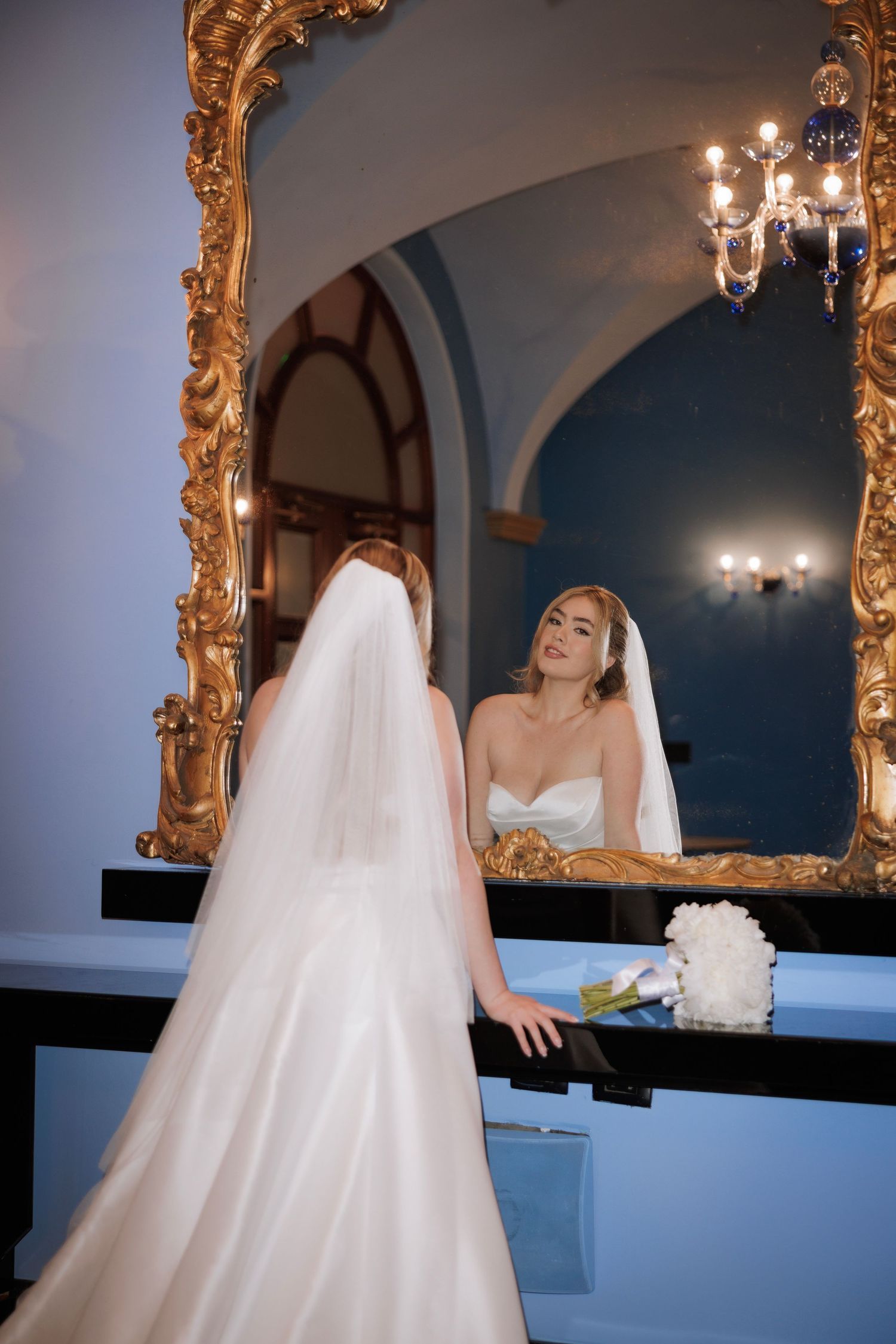 Wedding dress details captured in an ornate gold mirror with chandelier reflections.