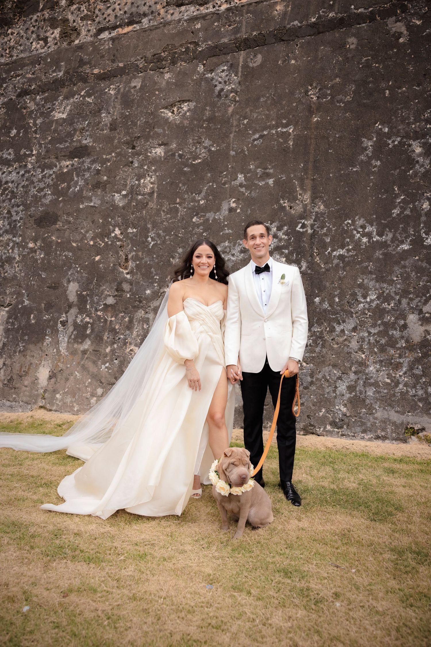 Wedding couple poses against weathered stone wall on grassy area.
