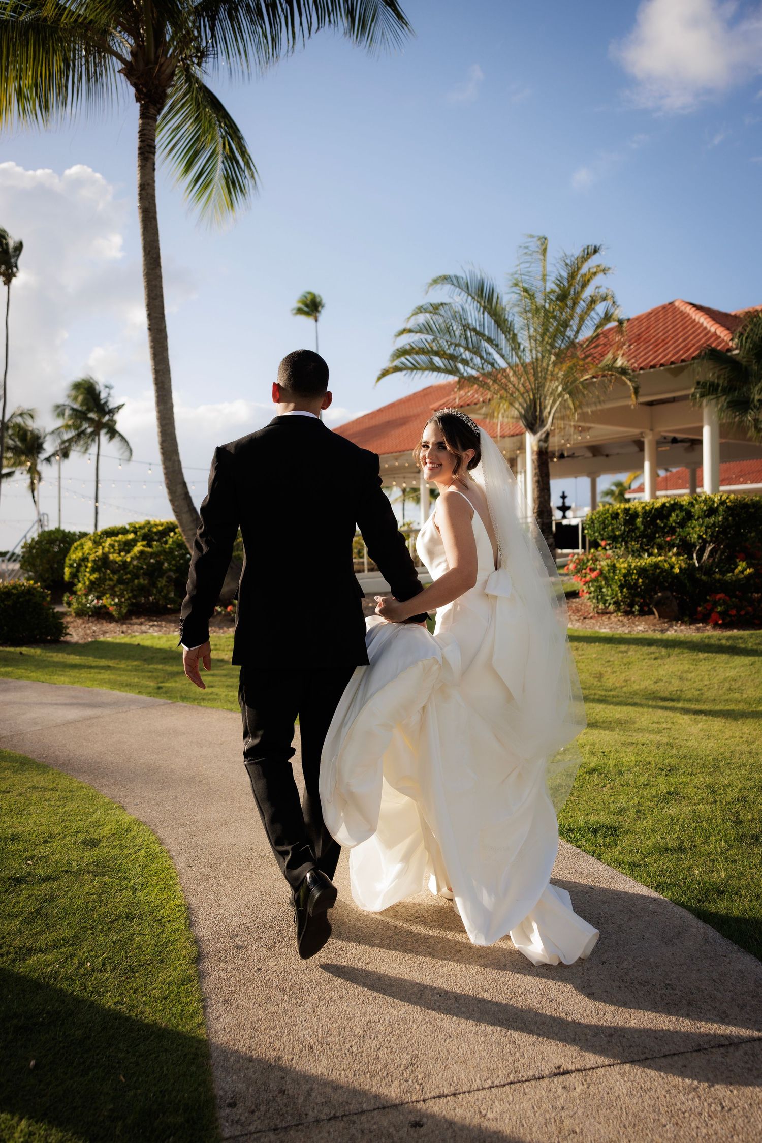 A couple in wedding attire walking hand in hand along a tropical palm-lined path at sunset.
