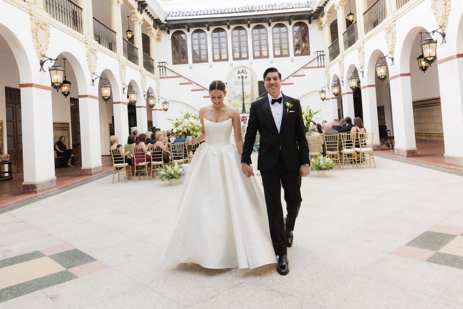 Elegant wedding couple walking through a Spanish colonial style courtyard with white columns and arches.