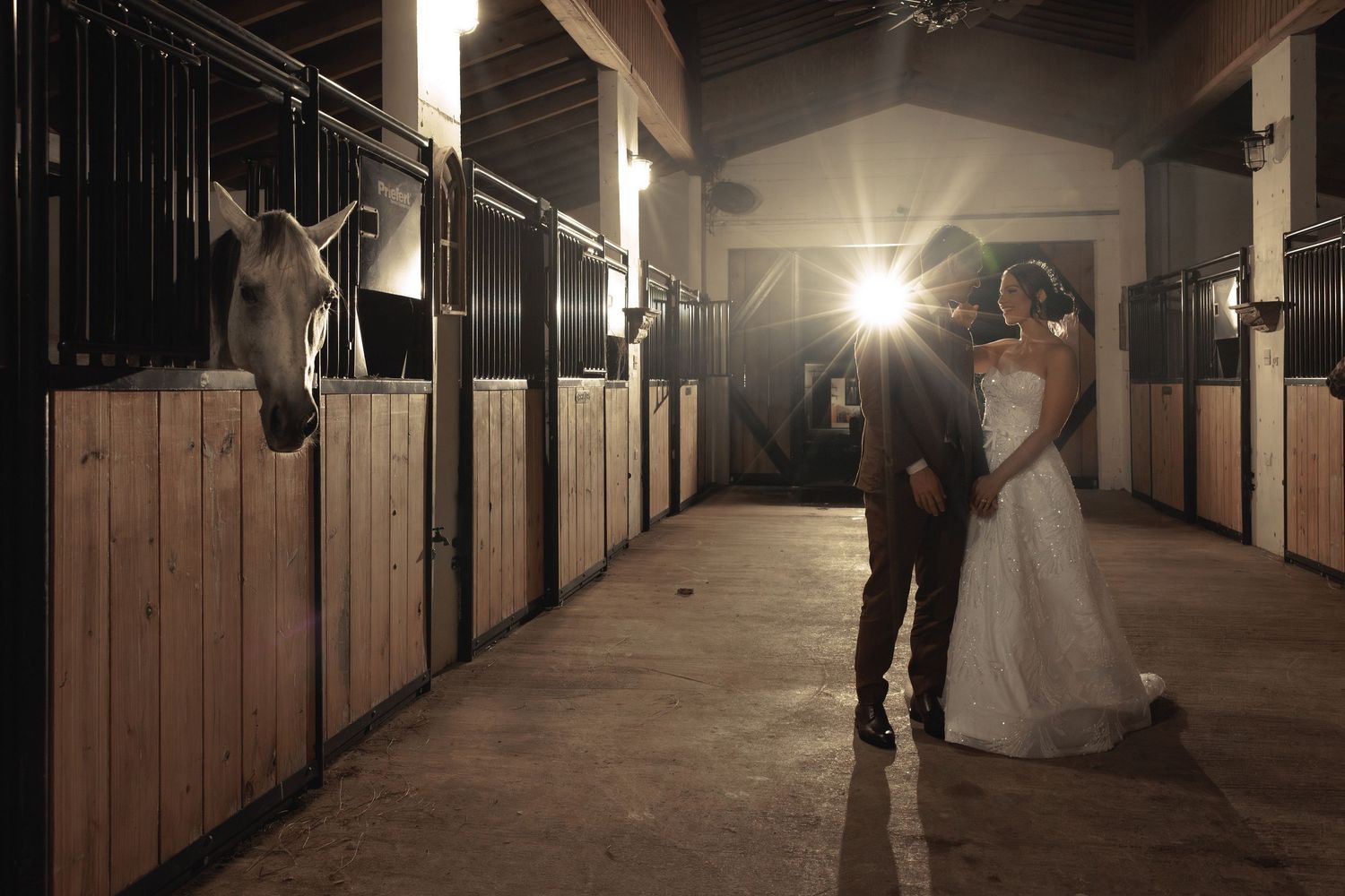 A silhouetted couple walks down a dimly lit barn hallway with wooden stalls and dramatic lighting.