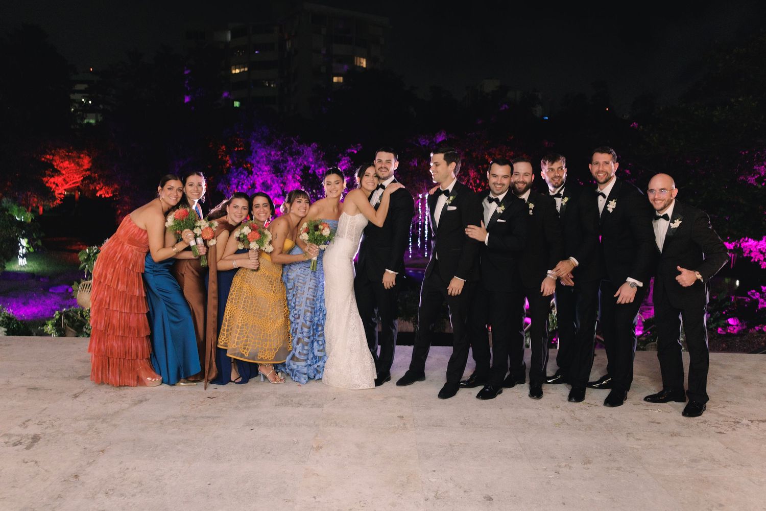 Wedding guests dance and celebrate on a dimly lit dance floor with colorful purple lighting in the background.