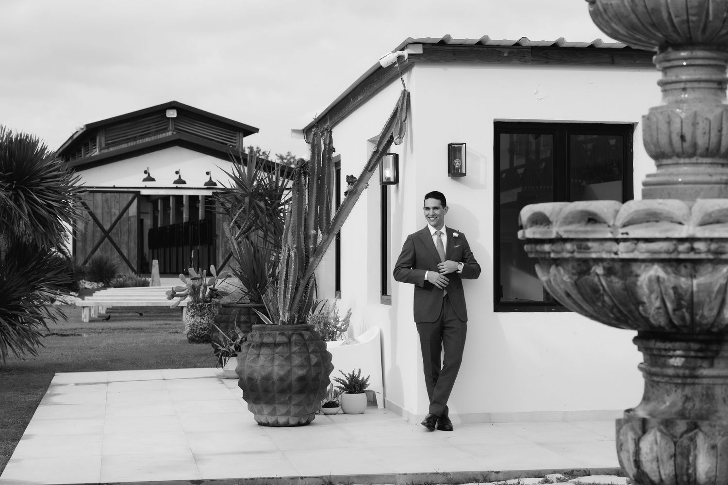 A person in a dark suit walks along a Spanish-style building with potted plants and a decorative fountain.