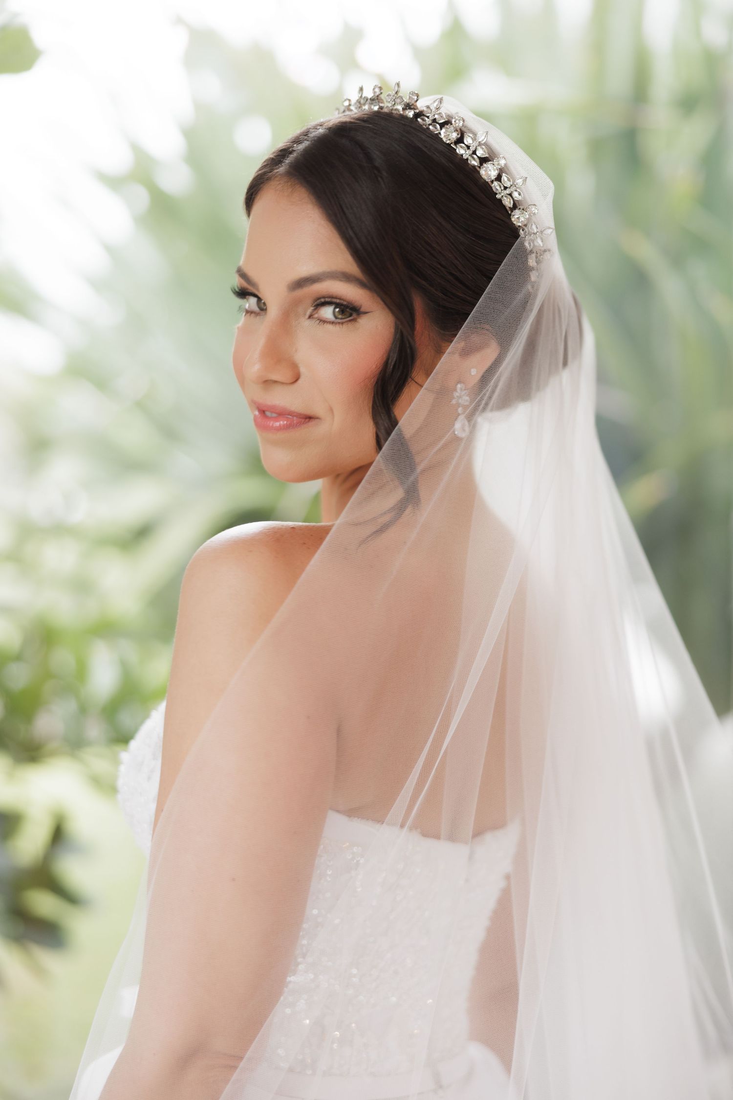A bride wearing an elegant crystal crown and delicate veil poses against a soft green outdoor backdrop.
