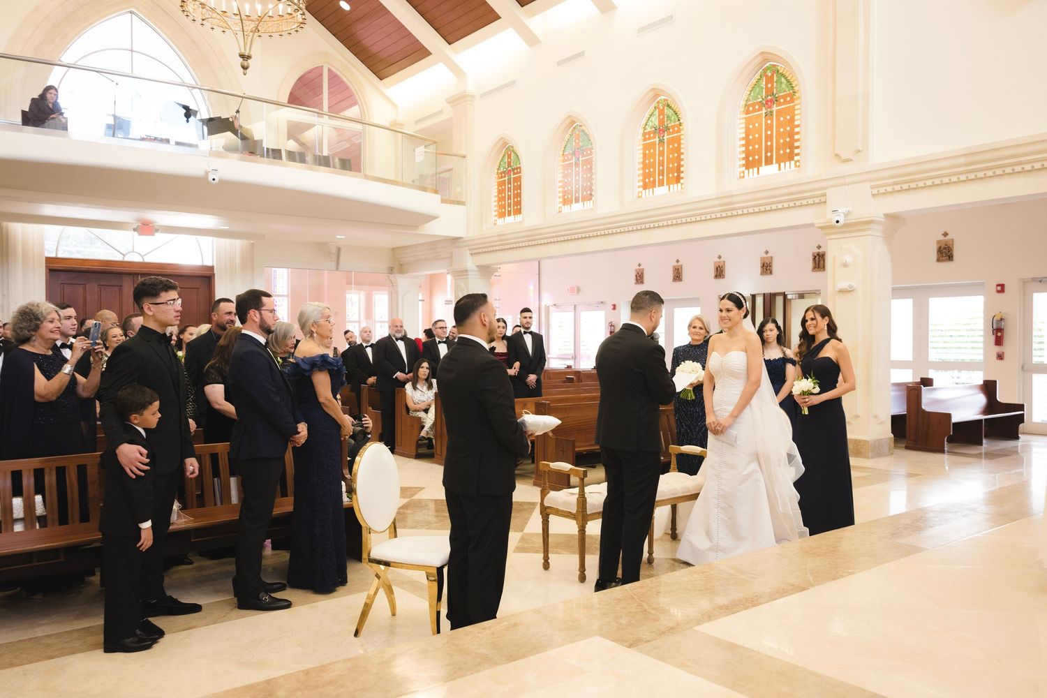 A joyous congregation rises from wooden pews in an elegant white church interior with arched stained glass windows.