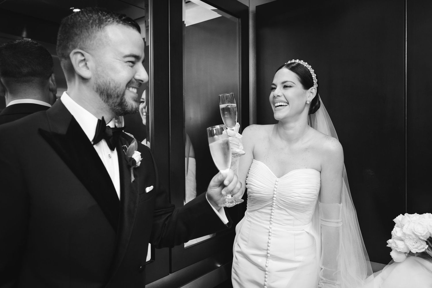 A joyful couple toasts with champagne flutes at their wedding celebration, captured in black and white.