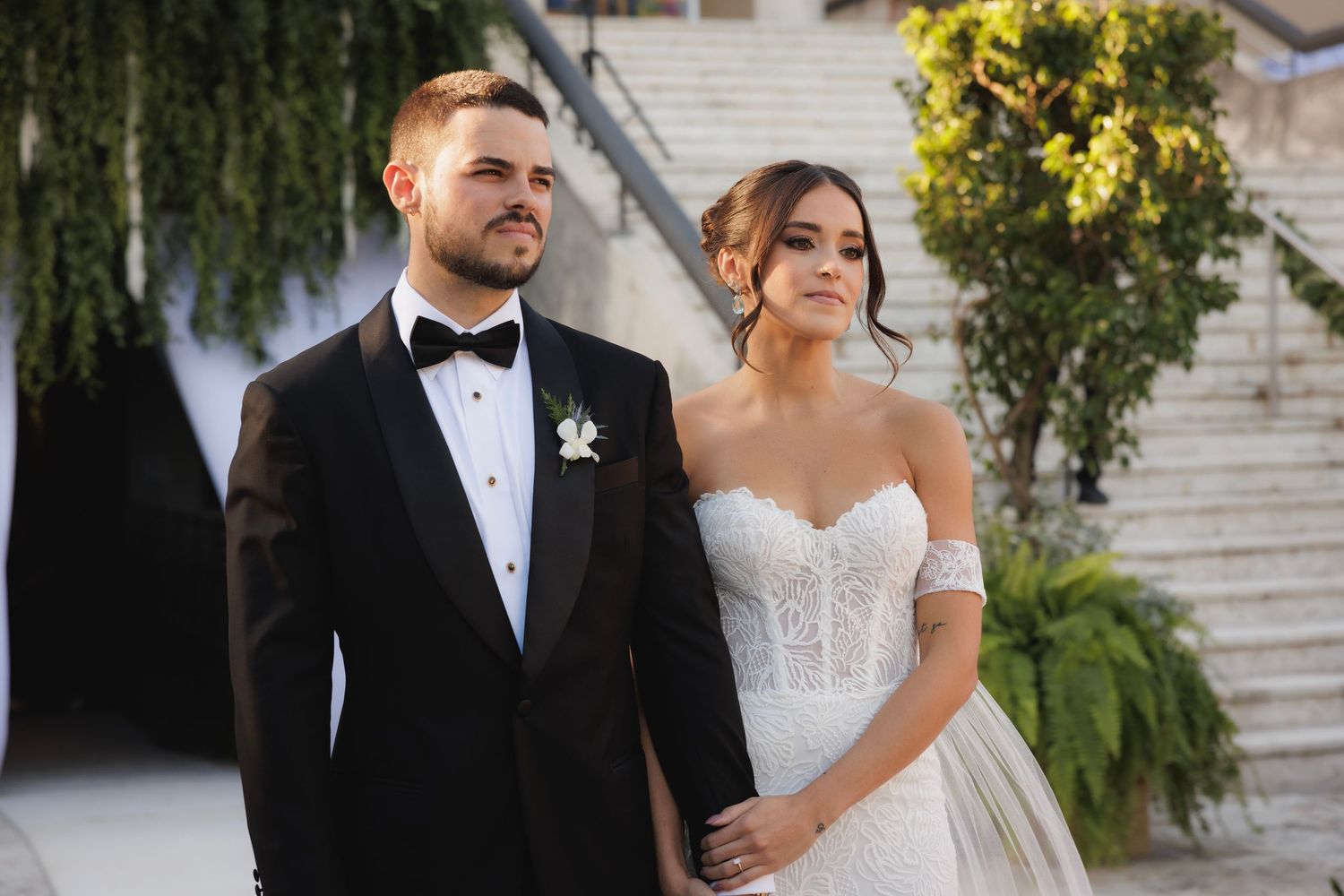 An elegant couple in formal wedding attire standing together at a stone venue with cascading greenery.