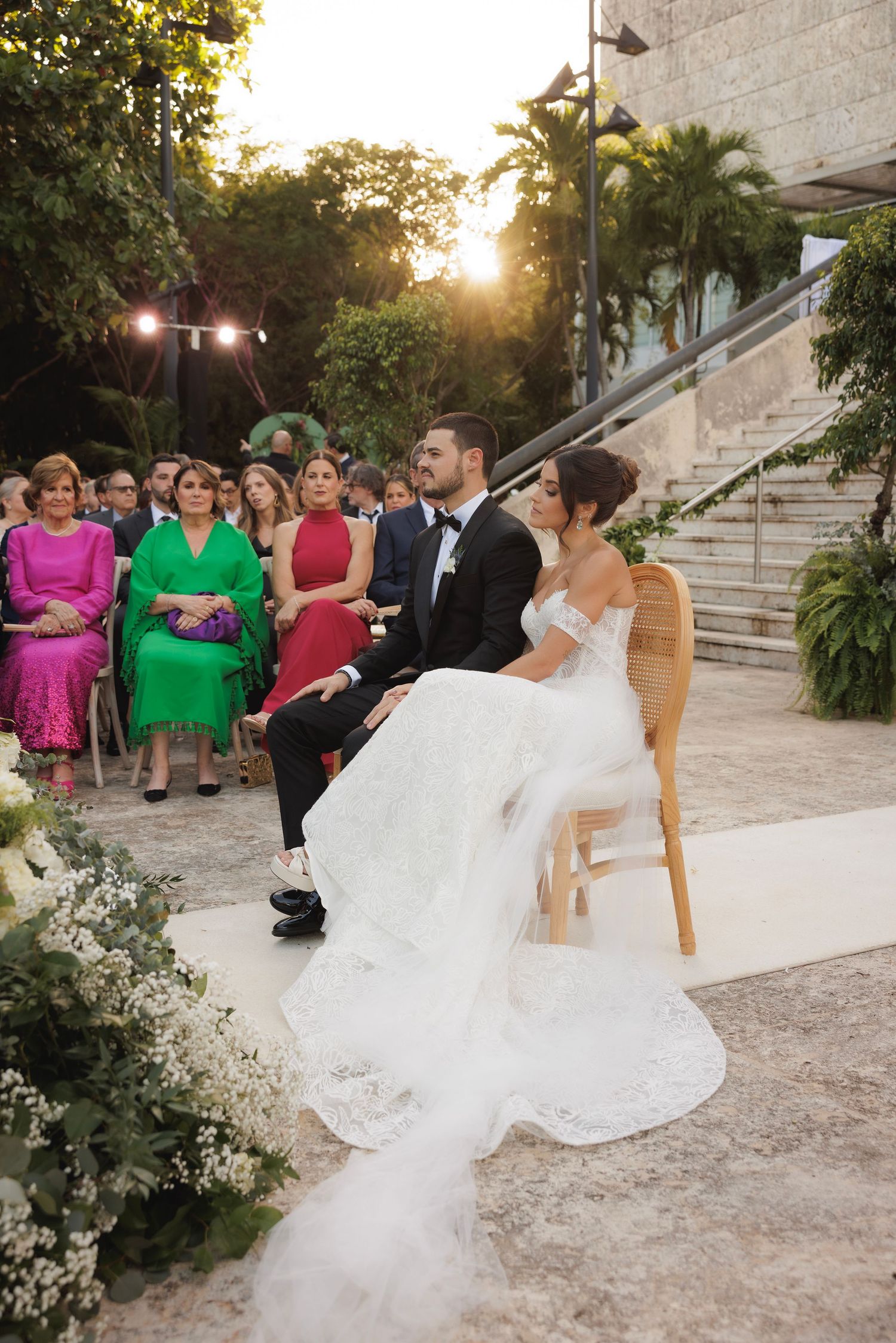 A sunset wedding ceremony with guests in vibrant attire watching the couple seated on wooden chairs.