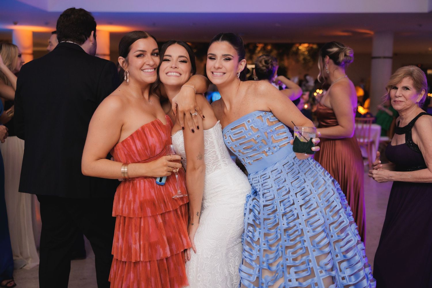 Wedding guests dance and socialize together at a reception with purple and pink lighting in the background.