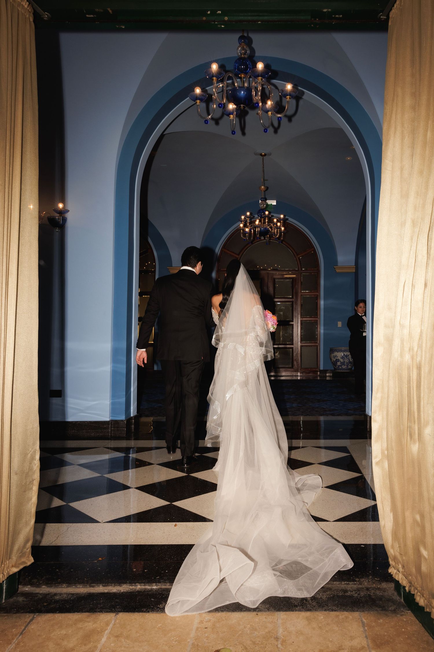 A couple enters an elegant hallway with checkered floors and blue arched ceilings adorned with crystal chandeliers.