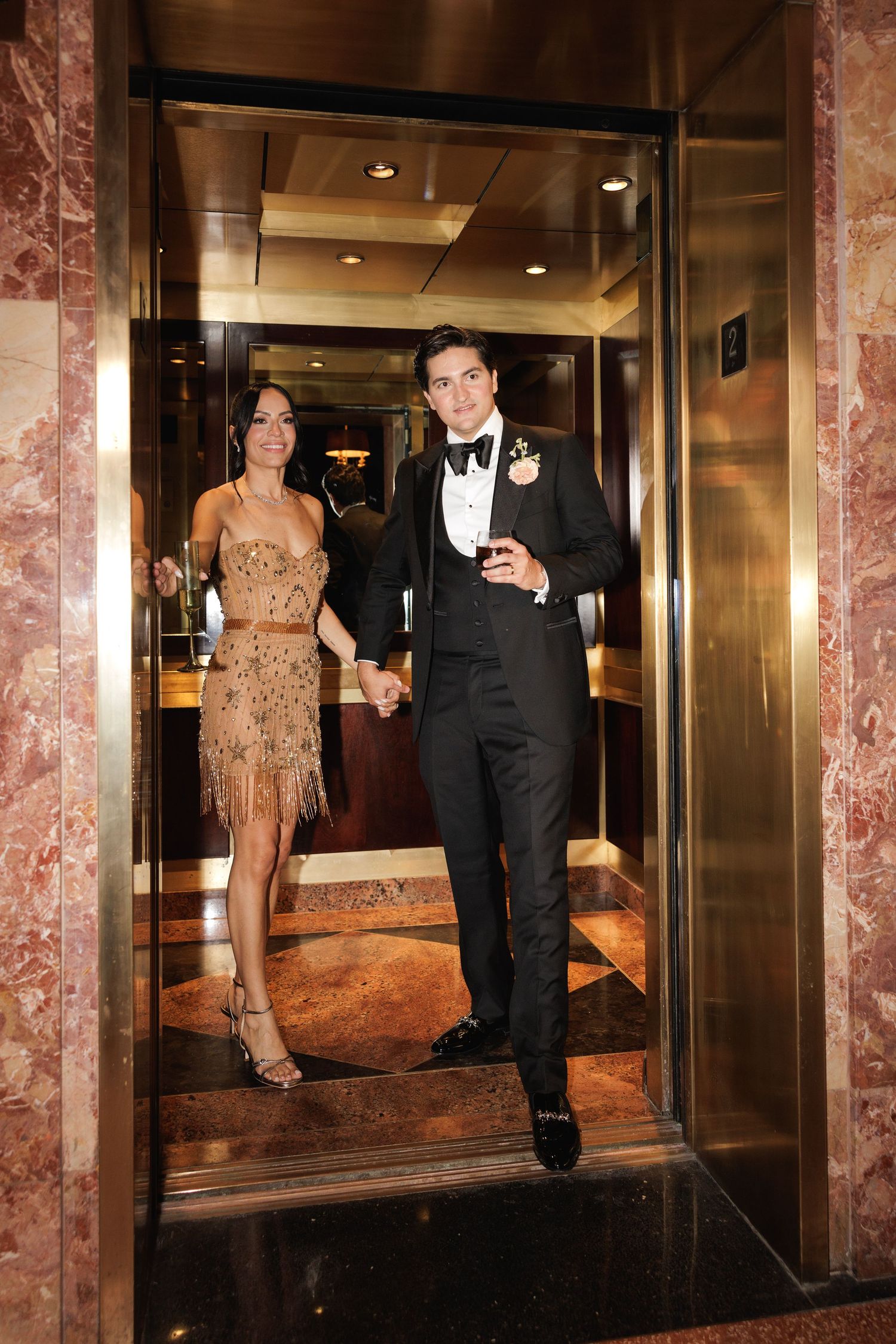 A couple in formal attire stands in an elegant hallway with marble columns and mirrored walls.