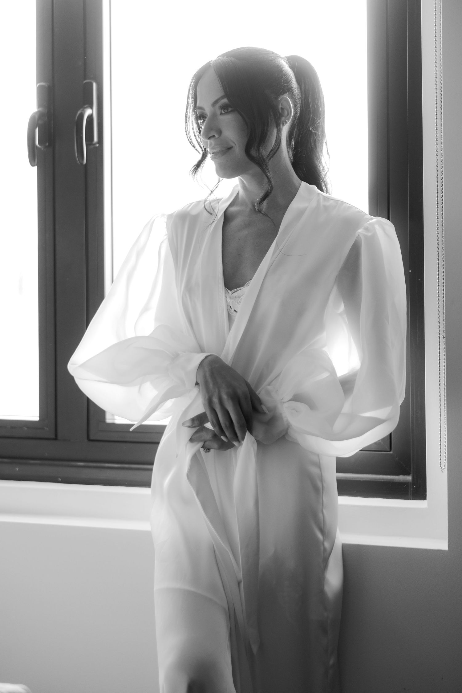 Artistic black and white portrait of a bride wearing a flowing white robe by a window.