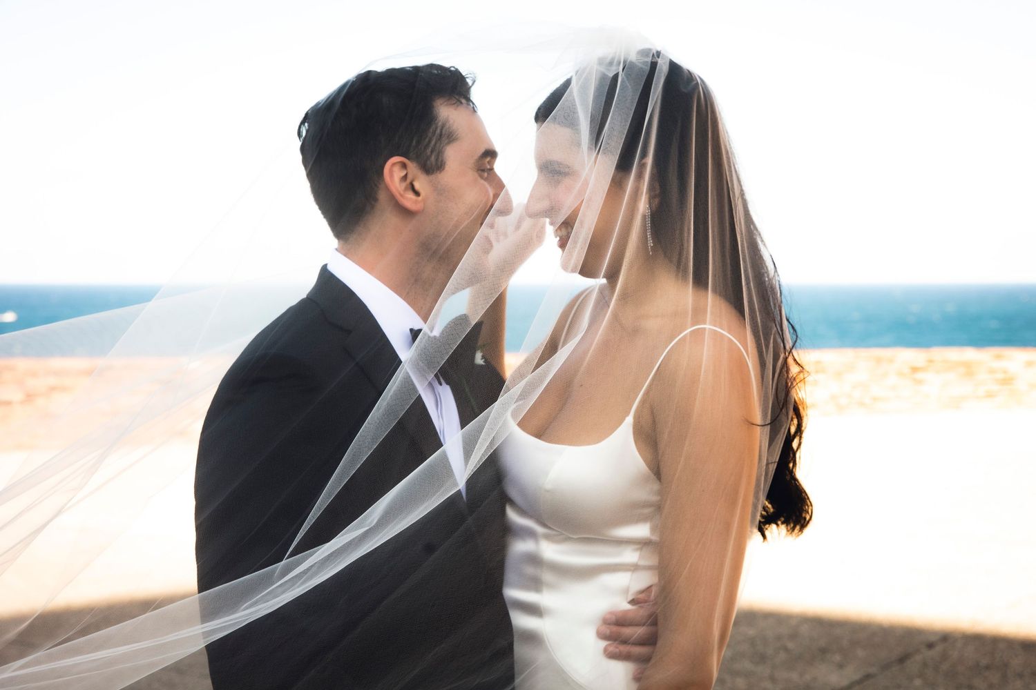 An intimate moment between newlyweds with a flowing veil against an ocean backdrop.