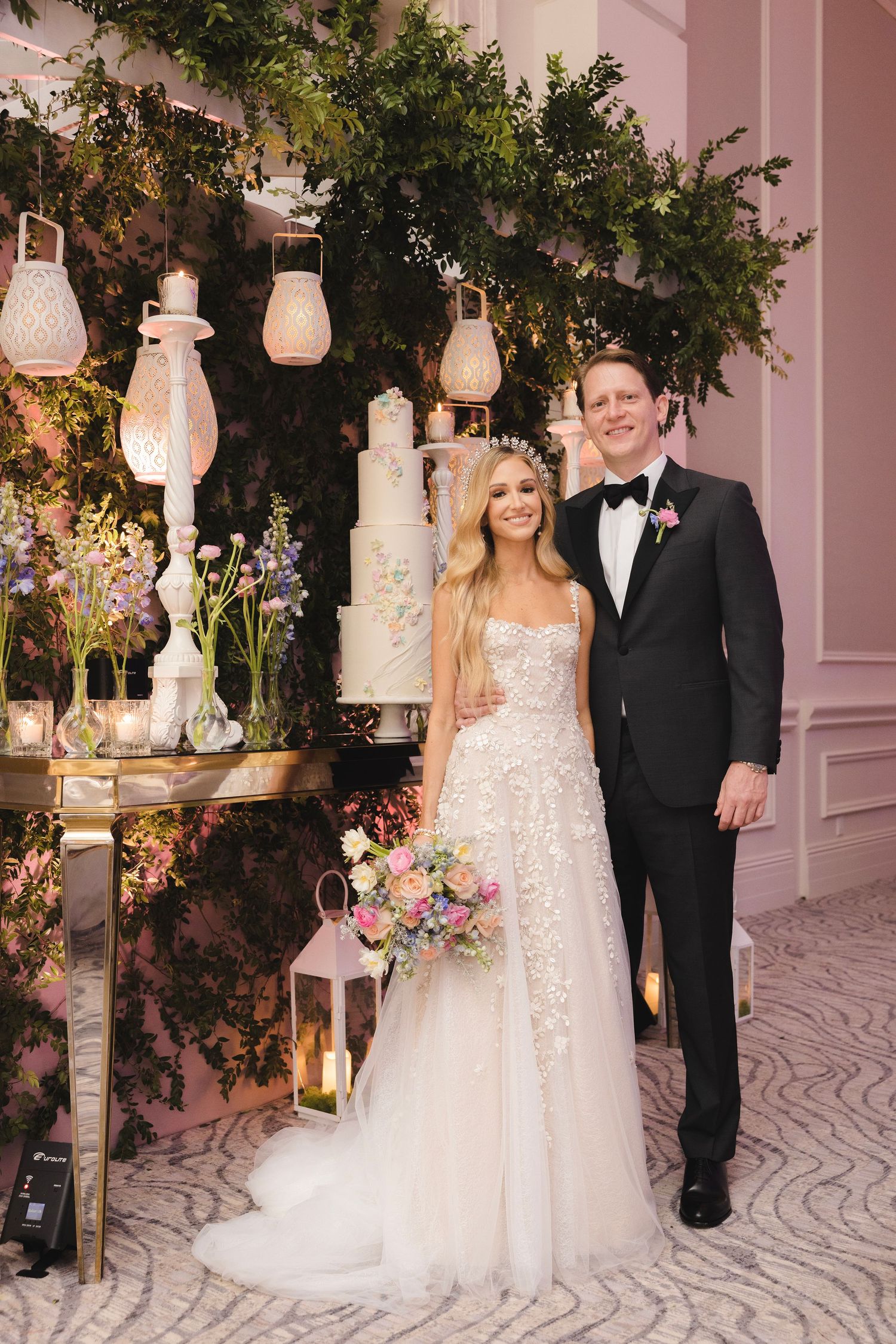 A glamorous wedding setup with a white tiered cake, hanging lanterns, and greenery against a pink wall.