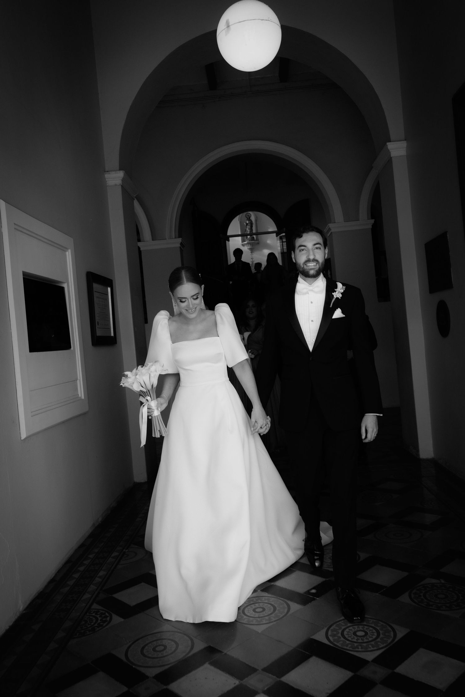 A black and white photo of a couple walking through an arched hallway in wedding attire.