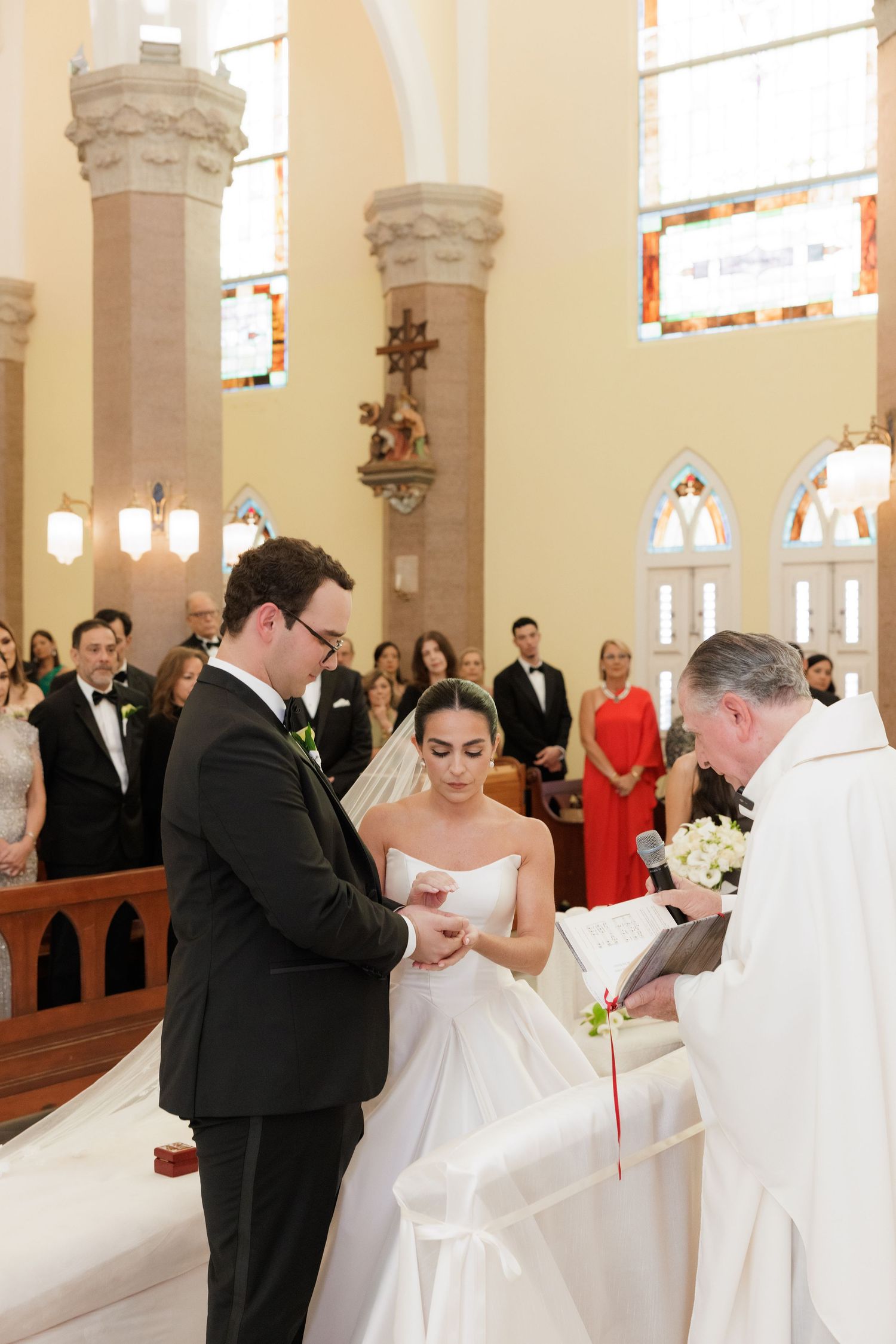 A wedding ceremony unfolds at the altar with the priest officiating as guests look on in a beautiful church setting.