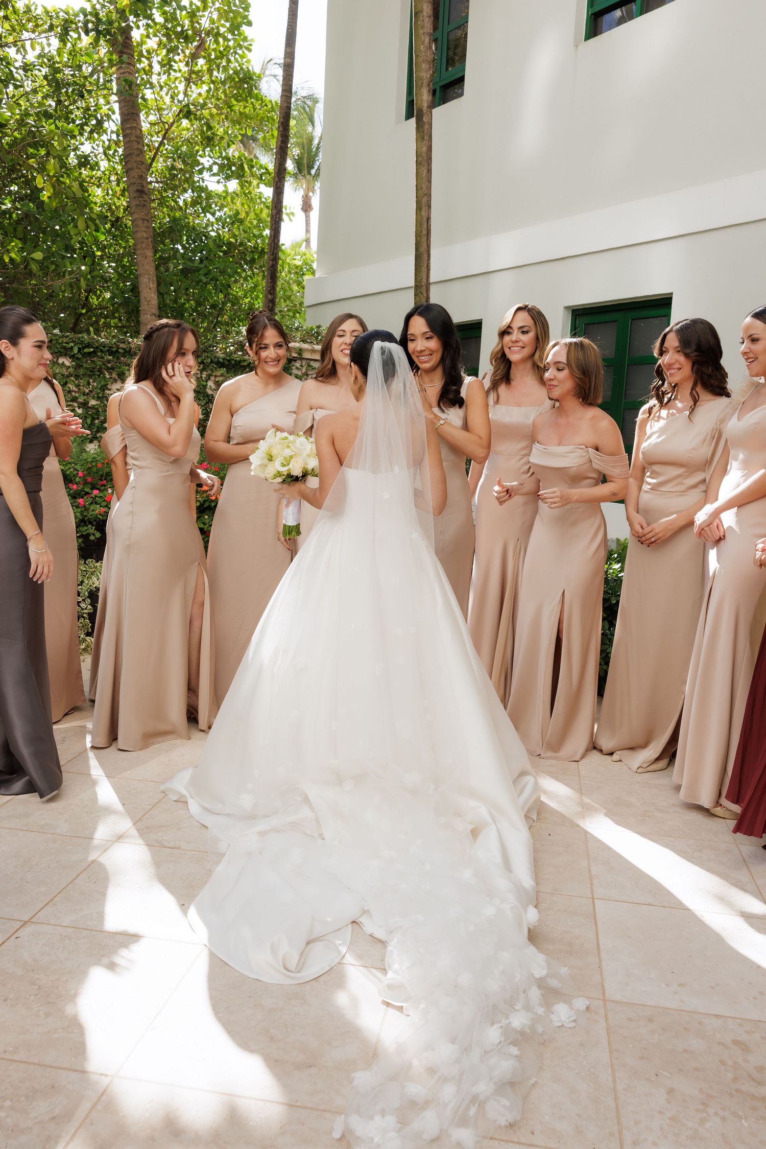A bridal party in blush pink bridesmaid dresses gathers around a bride in a white wedding gown with flowing train.