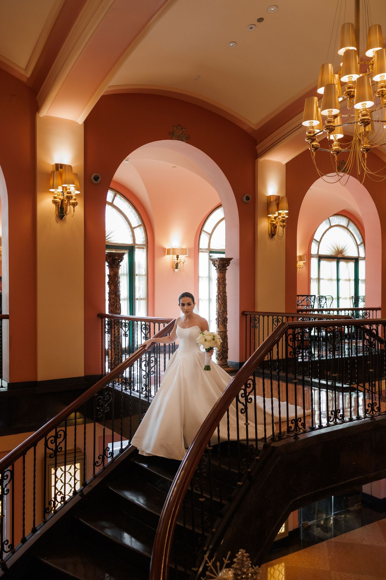 A bride in a classic white ballgown poses on a grand staircase with ornate chandeliers and coral-colored walls.