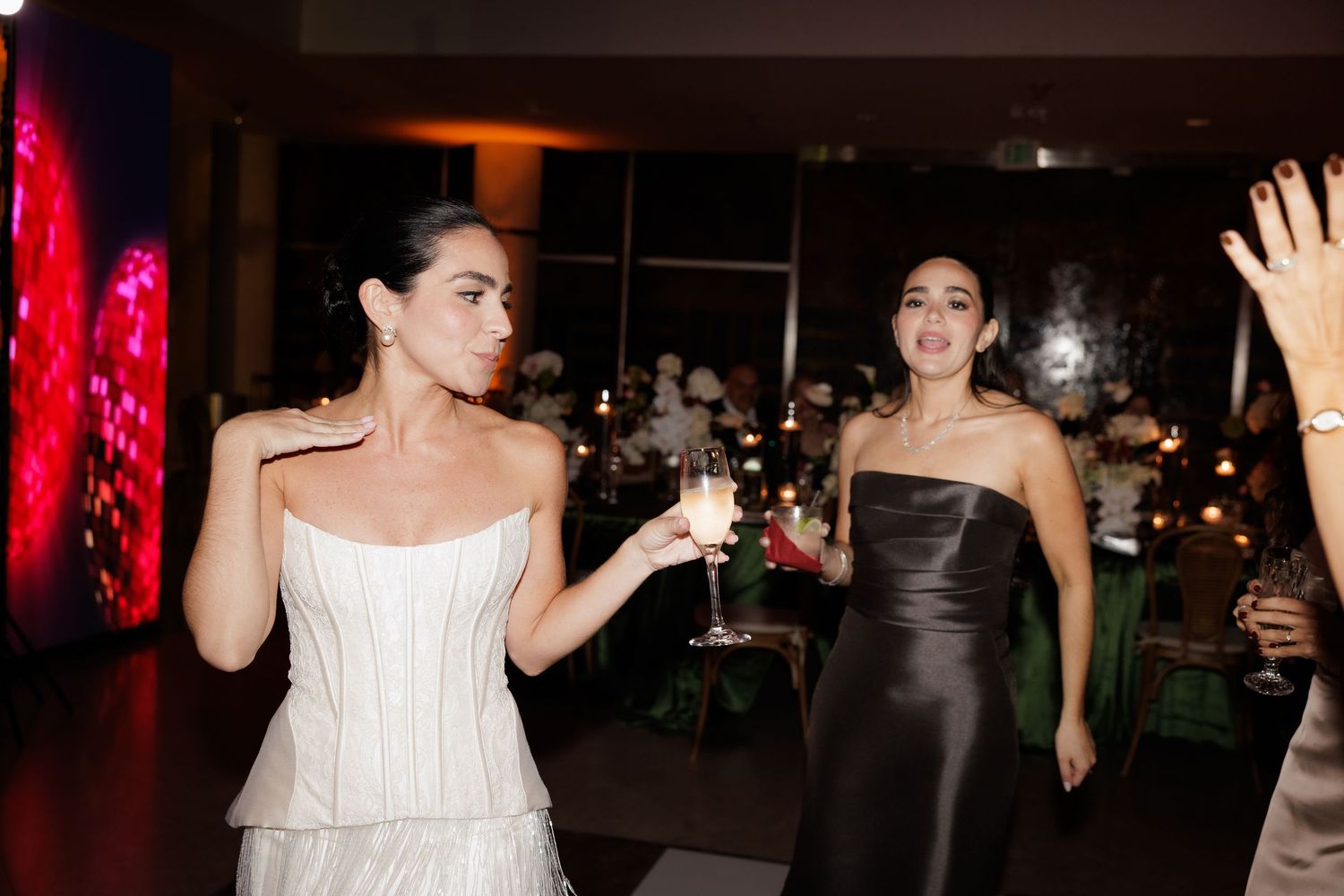 Two women in elegant dresses, one white and one black, raise their glasses in celebration at an evening event.